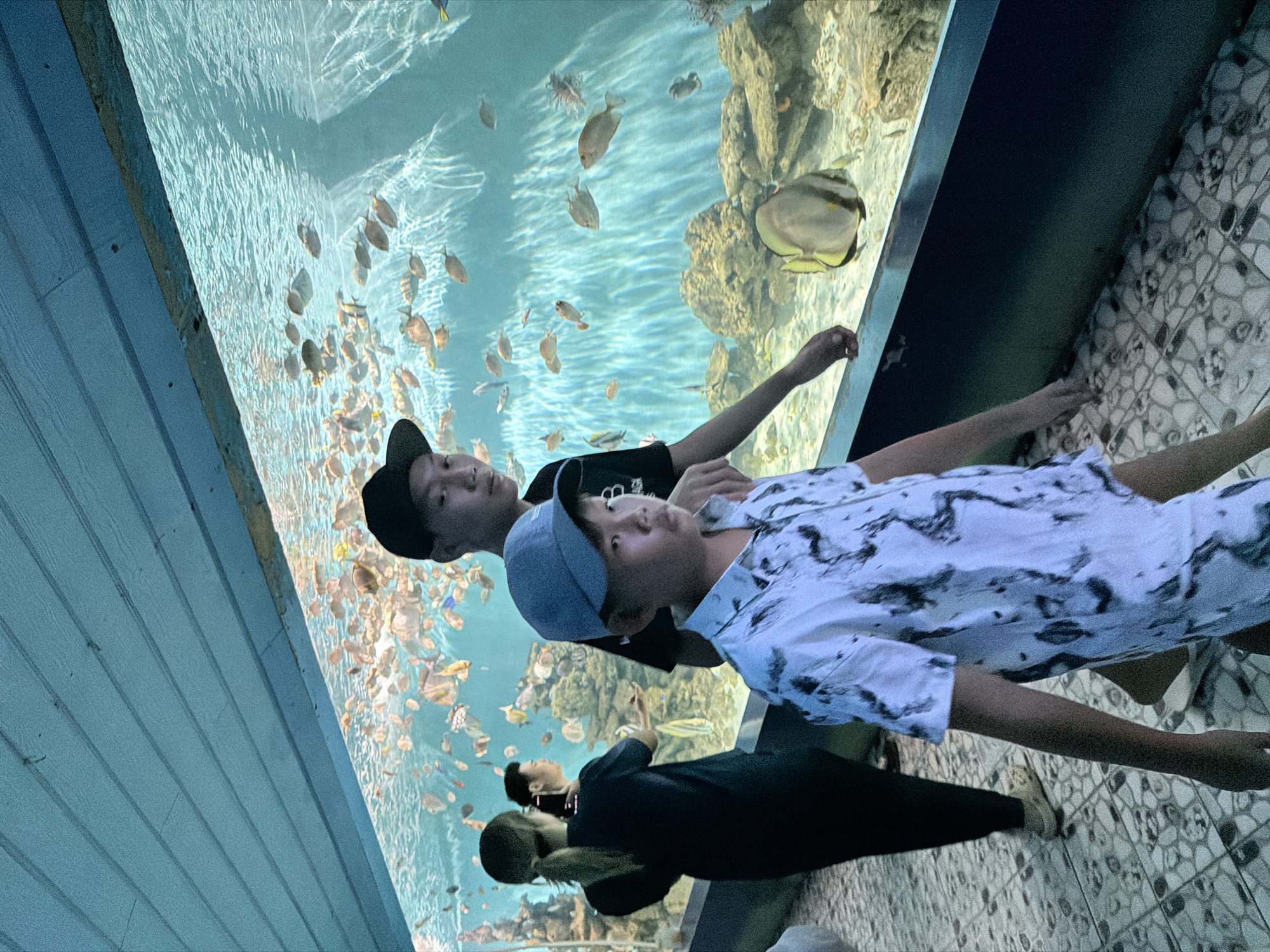 The image depicts an aquarium scene in Nha Trang, Vietnam. The foreground features two young males, one in a white patterned shirt and a blue baseball cap, and the other wearing a black t-shirt and a black baseball cap. They are positioned near a large aquarium tank filled with numerous fish and coral formations. In the background, two individuals, a female with her hair in a ponytail and a male, are observing the aquarium. The aquarium tank is situated beneath a light blue ceiling with visible wooden planks. The floor is tiled with a mosaic pattern. The overall environment suggests a public aquarium attraction.