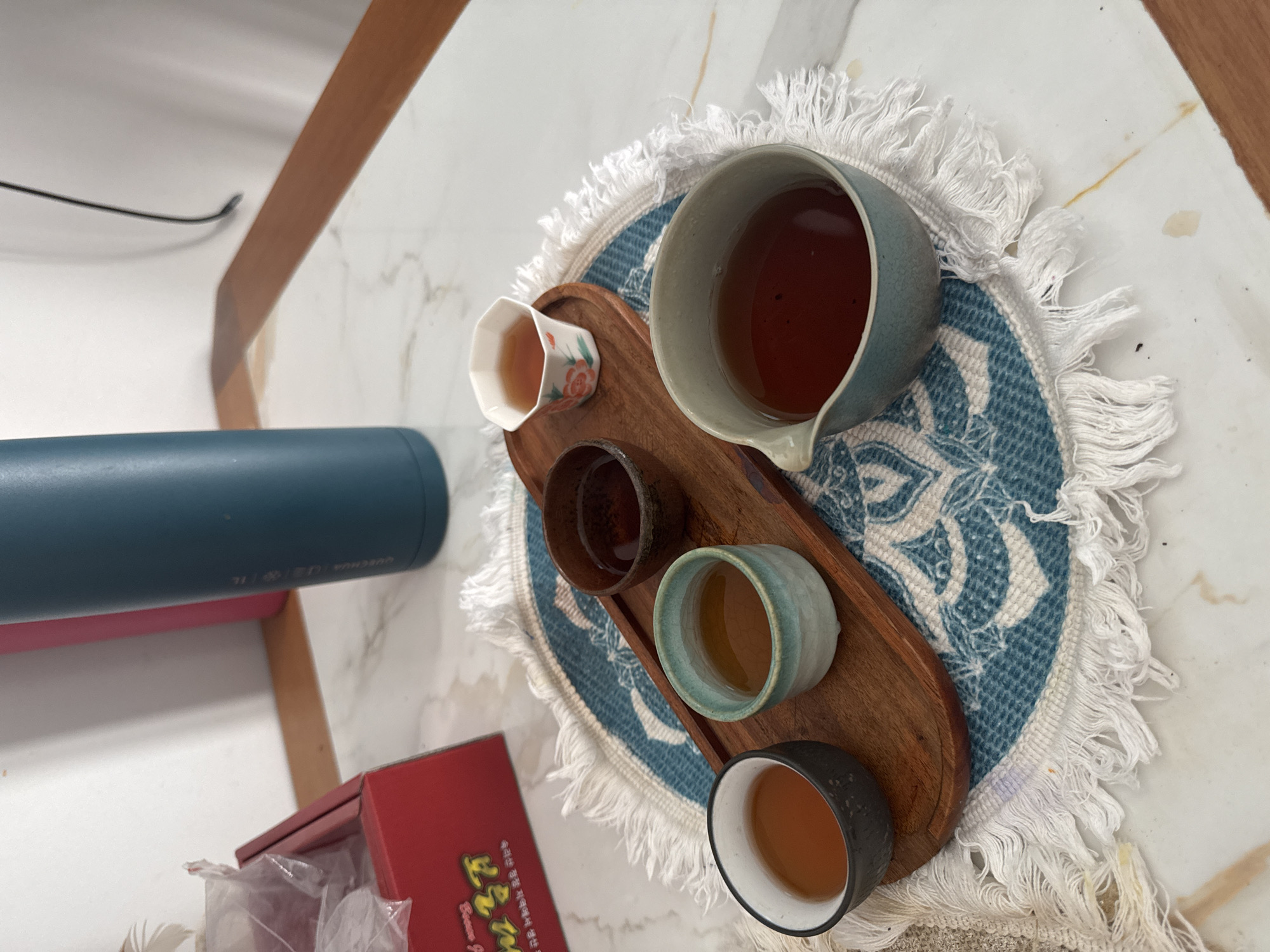 A top-down shot shows a tea set placed on a blue and white patterned circular placemat with fringe. The tea set comprises a spout cup and four teacups, all filled with tea. They rest on a wooden tray. To the left of the tray is a blue thermos and a red box. The scene appears to be set indoors, possibly on a table in a private residence. Lâm Viên - Đà Lạt, Vietnam is not visibly discernible.