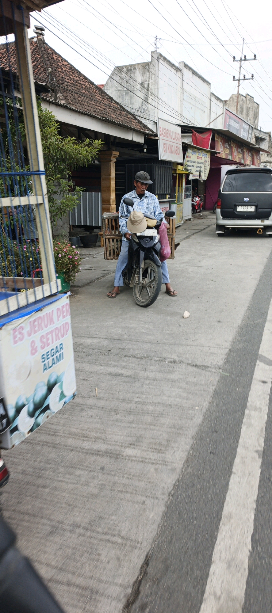 In Kasiyan, Indonesia, a man sits astride a motorcycle on a paved road. He is wearing a blue and white patterned long-sleeved shirt, light blue jeans, and flip-flops. A brown hat rests on the handlebars of the motorcycle, and a pink plastic bag hangs from the left handlebar. The man appears to be smoking a cigarette. Behind him, a cart with a blue and white sign reads "ES JERUK PER & SETRUP SEGAR ALAMI". The background shows buildings with tiled roofs and various signs, including one that says "BASRENG LUENGKING BY KING ABDI". A silver van with a black license plate is parked to the right of the motorcycle. The scene depicts a street-side vendor in a typical Indonesian setting.