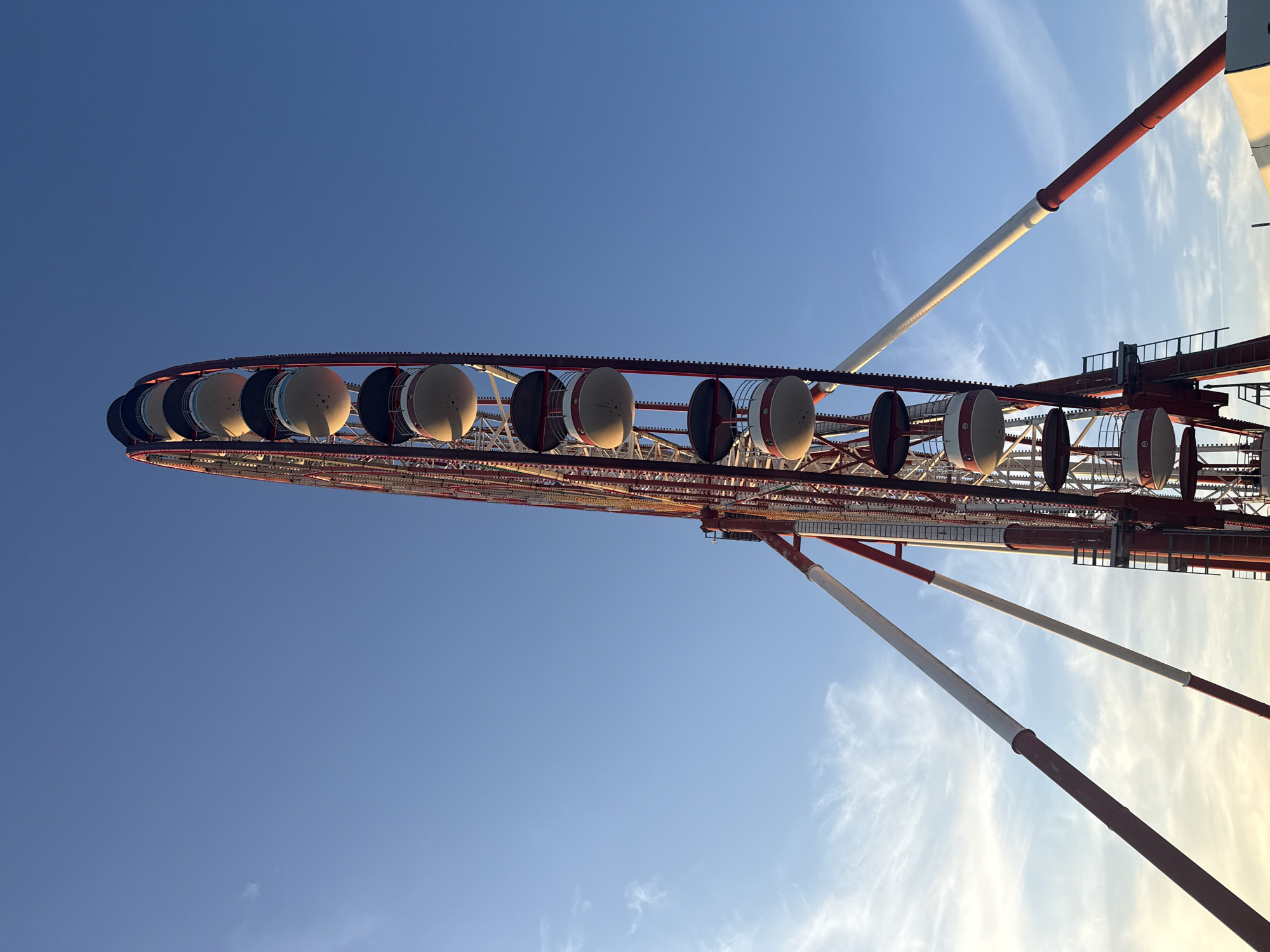 A large Ferris wheel, identified as located in Batumi, Georgia, is presented from a low-angle perspective, emphasizing its height against a clear to partly cloudy sky. The structure of the Ferris wheel is predominantly red and white, with thick, angled support beams painted in a red and white striped pattern. The main wheel mechanism, visible on the left side, features a red frame and numerous internal white or silver bracing elements. Multiple enclosed gondolas, cylindrical or capsule-shaped, are attached to the wheel's circumference. These gondolas are painted white with a red band near the bottom and feature dark, possibly black or dark blue, canopies or roofs. Sunlight illuminates the left sides of the gondolas, creating shadows on their right sides. The sky transitions from a lighter, yellowish-blue on the lower left horizon, indicative of late afternoon or early evening light, to a deeper, darker blue towards the upper right. Wispy white clouds are present in the lighter portion of the sky on the left. No people or other city elements are visible, focusing solely on the Ferris wheel and the sky.