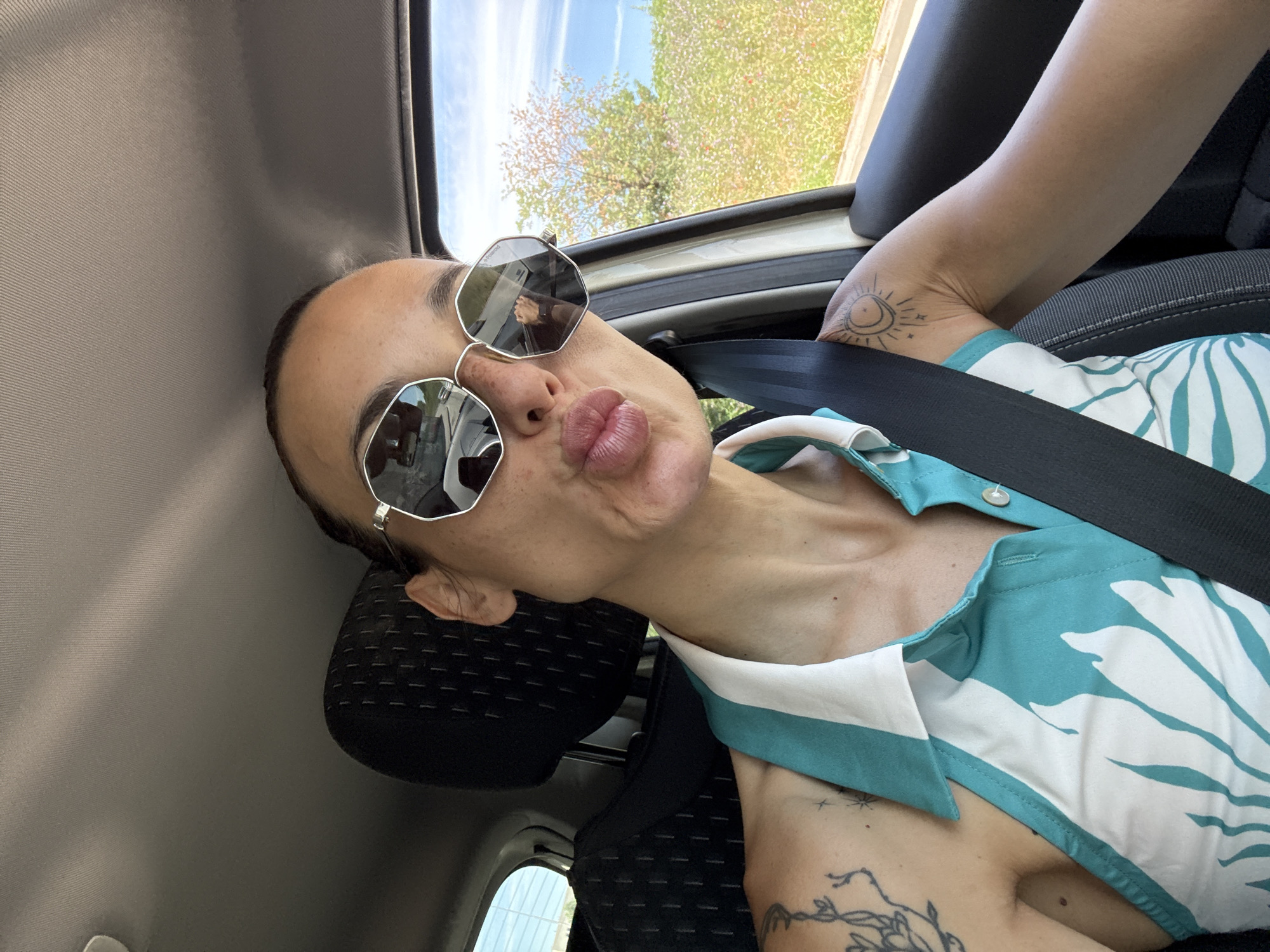 A woman with sunglasses and full lips is taking a selfie inside a car. She is wearing a teal and white patterned top and has tattoos on her left arm and shoulder. The car's interior is visible, including the headrest and seatbelt. Outside the car window, a field of wildflowers and trees can be seen under a blue sky. The exact location in Pleven, Bulgaria is not discernible from the image.