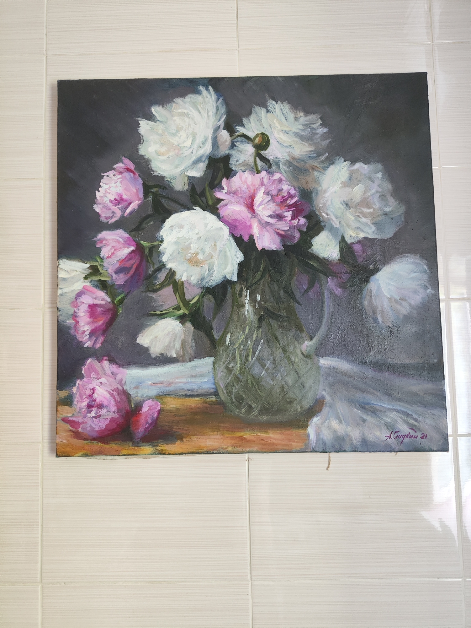 A framed oil painting depicts a bouquet of peonies in a clear glass vase. The vase is placed on a wooden surface, with a white cloth draped to the right. Some peonies are white, while others are pink. One pink peony lies on the surface in front of the vase, and another bud is visible to its right. The background is a dark, textured gray. The artist's signature, "А. Смугуин 21", is visible in the lower right corner of the painting. The painting is hanging on a tiled wall, with the tiles having a light, textured finish. There are no people or animals in the image. The painting is likely displayed indoors, and its location within the city of Kharkiv, Ukraine, is not determinable from the image alone.