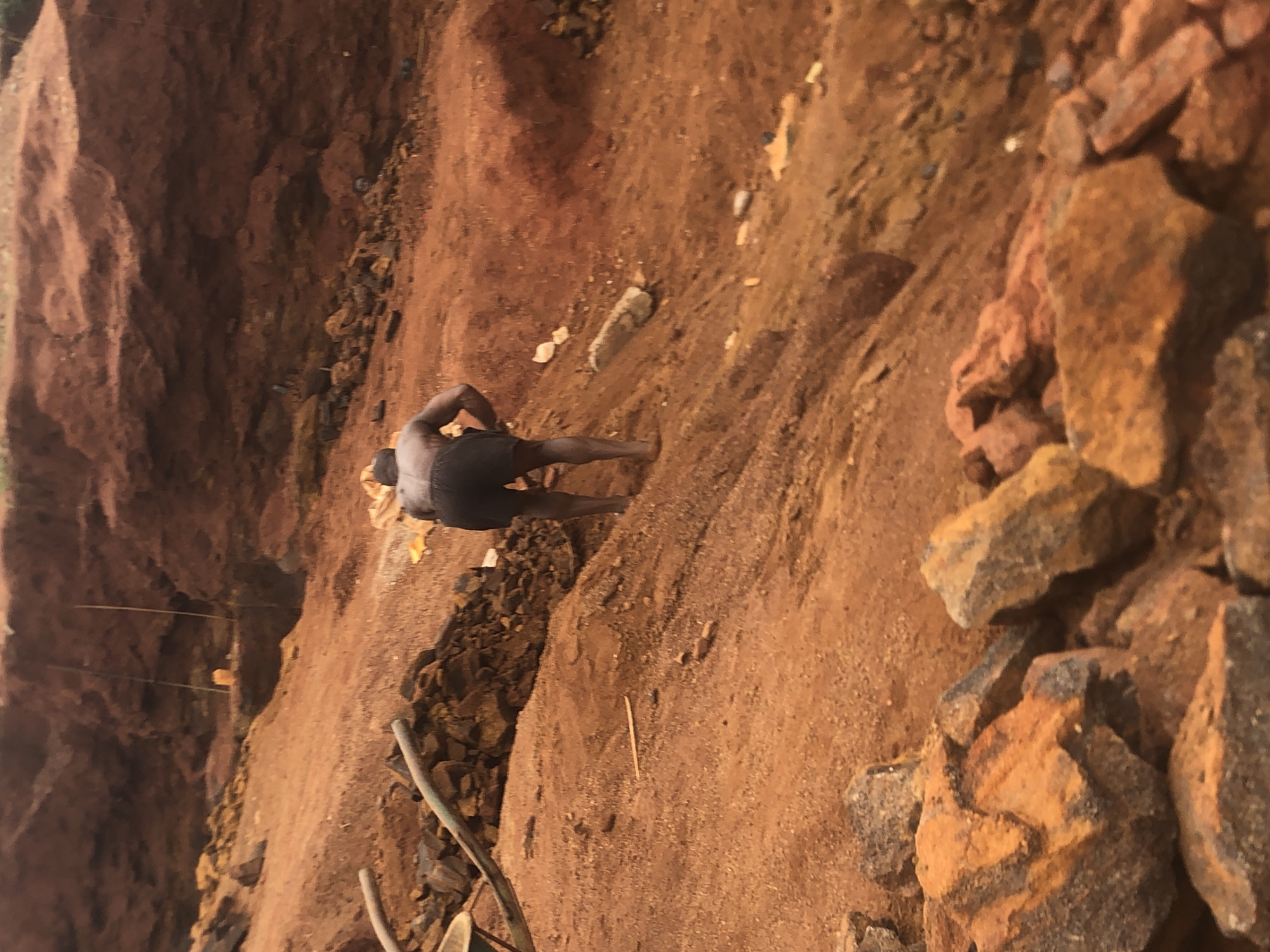 The image, taken in Ukwu-Abwa, Nigeria, captures an outdoor scene at what appears to be an excavation or quarry site. The dominant features are reddish-brown earth and numerous angular rocks, characteristic of lateritic soil or iron-rich deposits.
A single individual, a person with dark skin, is seen from the back in the lower-left portion of the frame. They are wearing dark shorts or underwear and appear to be barefoot, standing on the uneven, rocky ground. Their posture is slightly hunched, suggesting a moment of rest or observation during physical labor.
The background consists of steep, exposed earth banks, while the foreground and mid-ground are covered with loose dirt, small stones, and larger rock fragments. A few thin sticks or poles are visible on the ground. The lighting indicates it is daytime, with bright but possibly diffused light, as there are no distinct harsh shadows. The ground appears dry, suggesting fair weather. The scene conveys a sense of rugged, manual work in an open, undeveloped environment. There is no visible text in the image.
