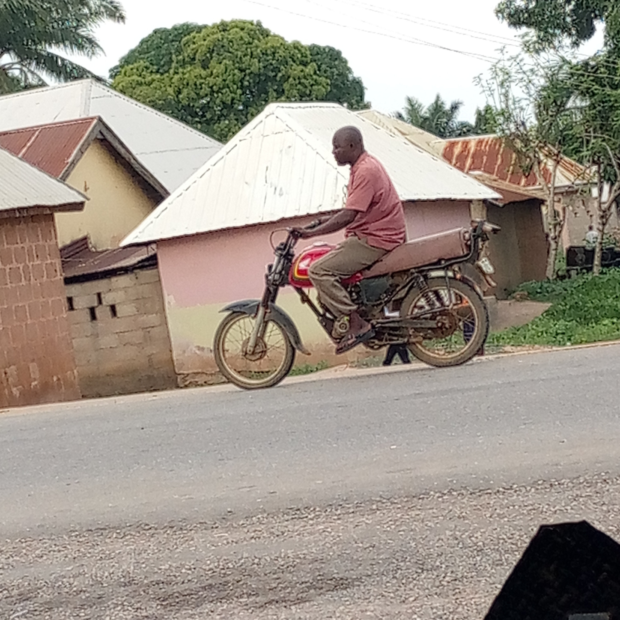 The image depicts a man riding a motorcycle on a paved road in Mararaba, Nigeria. The man is wearing a reddish-brown shirt and khaki pants, and is likely traveling at a slow speed.
The background shows a cluster of buildings with corrugated metal roofs, some painted white and others red. There are green trees and shrubs interspersed among the buildings. A short brick wall is visible on the left side of the frame. The sky is overcast. The road appears to be paved. A small amount of the dashboard of a vehicle is visible in the bottom of the frame, suggesting the image was taken from inside a car.