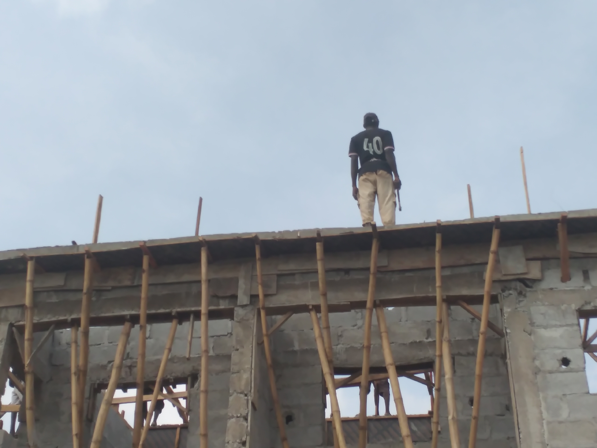 A construction worker stands on the top floor of a building under construction, looking out at the sky. The worker, wearing a black shirt with "40" printed on the back and light-colored pants, holds a tool in their right hand. The building appears to be made of concrete blocks and is supported by wooden scaffolding. The sky is overcast.