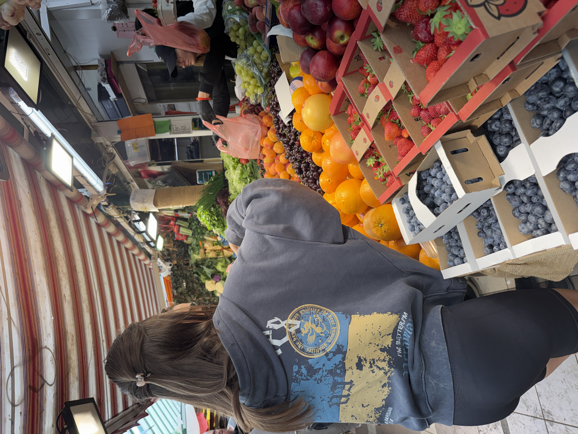 A medium shot, taken from slightly above, captures a person with long brown hair and wearing a gray hoodie with an emblem, standing at a vibrant fruit and vegetable stand. The person is facing away from the camera, their back towards the viewer. The emblem on the hoodie appears to have a circular design with an image of a griffin and lettering inside, as well as a blue and yellow backdrop.
The stand is overflowing with fresh produce, including blueberries in white cartons, strawberries in cardboard containers, oranges, apples, and other fruits and vegetables. The fruits and vegetables are arranged in an organized display, creating a colorful and appealing sight.
The background reveals a glimpse of the marketplace setting, with striped awnings, lighting fixtures, and other vendors. The lighting is bright, highlighting the freshness of the produce and the overall lively atmosphere of the market.