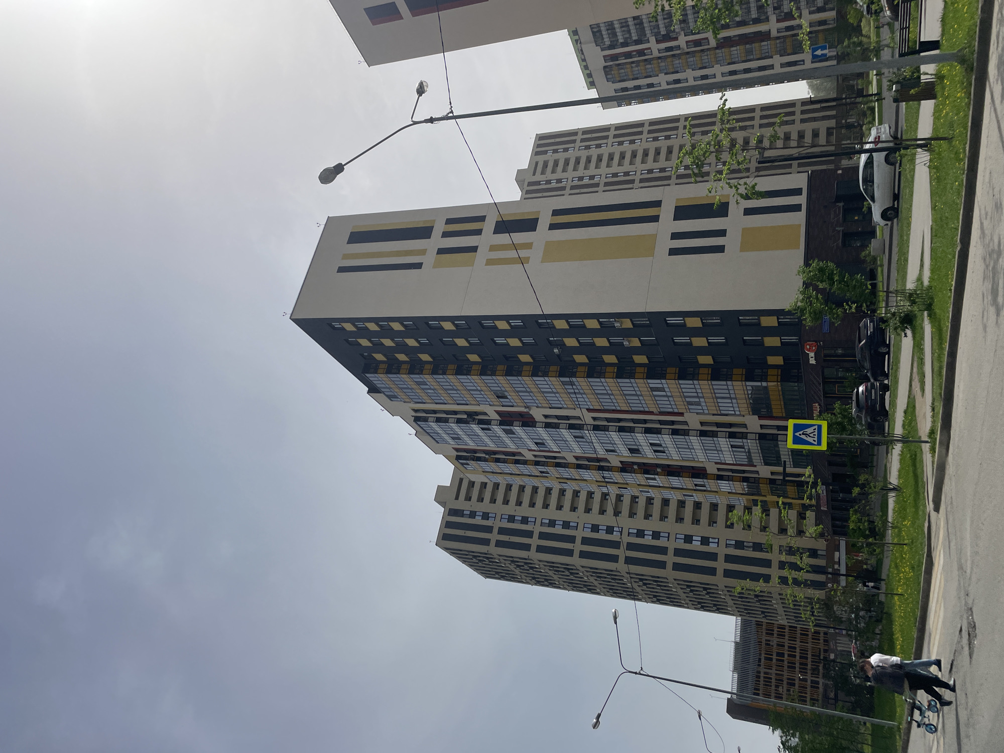 The image shows a street scene in Kazan, Russia, with a view of several tall apartment buildings. The buildings feature a modern architectural design with horizontal stripes of yellow and black on their facades. A street sign with a pedestrian crossing symbol is visible. There are a few cars parked along the side of the road. A person pushing a stroller walks along the sidewalk. Streetlights are present. The sky is overcast.