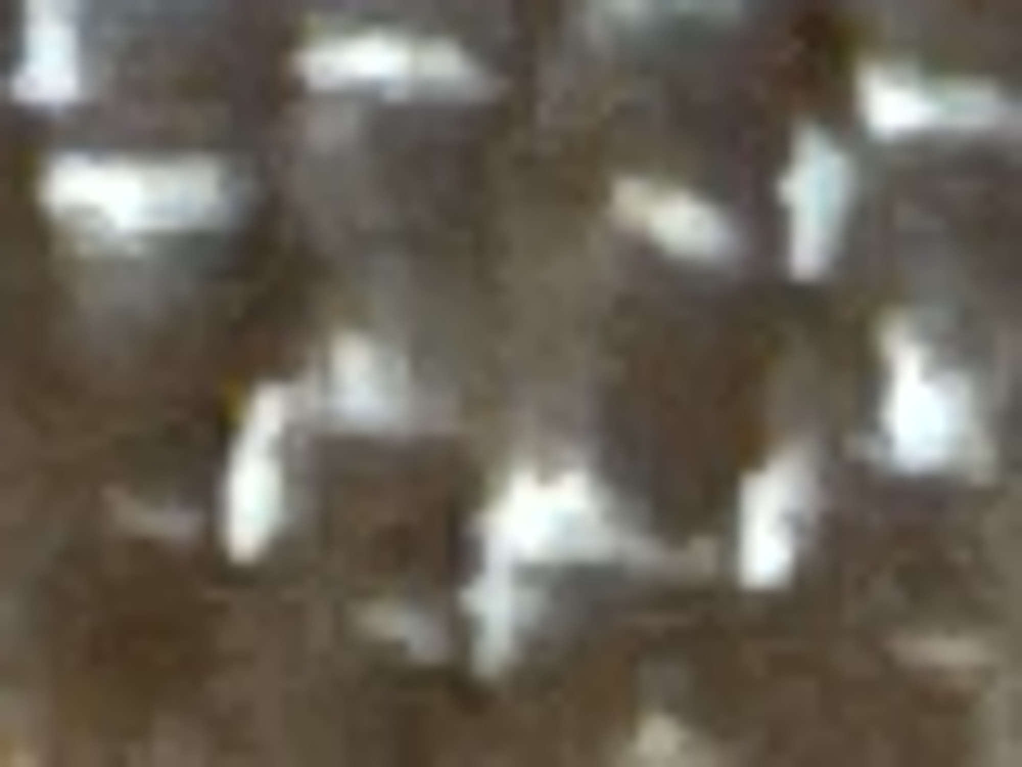 The image shows a close-up, pixelated view of a textured surface. The texture appears to be woven, with alternating dark and light strands creating a grid-like pattern. The colors are muted, primarily shades of brown and white, suggesting a material like burlap, coarse fabric, or possibly a carbon fiber weave. The lighting creates highlights on the lighter strands, emphasizing the texture. The image is too low-resolution to discern any specific details about its location within Navbahor, Uzbekistan, or to identify any subjects, objects, actions, interactions, or environmental features beyond the textural surface itself.