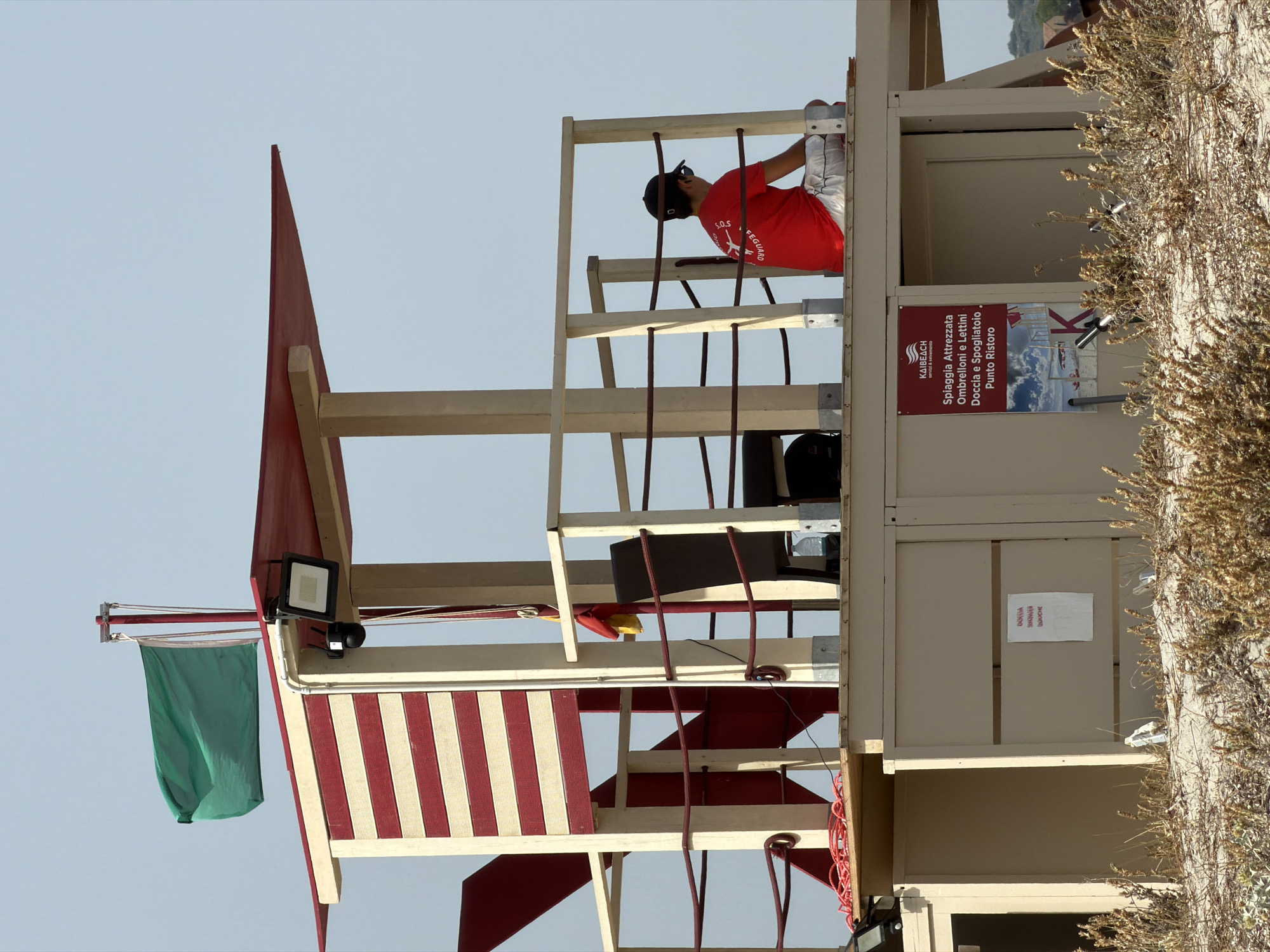 The image shows a lifeguard tower on a beach in Olbia, Italy. A lifeguard wearing a red shirt and white shorts is seated on the platform of the tower, facing away from the camera. A green flag is flying from the top of the tower. The tower has red and white striped panels on its sides. There are also signs on the lower part of the tower, one with Italian text that translates to "Equipped beach Umbrellas and Sunbeds Showers and Changing Rooms Refreshment Point." The ground in front of the tower is covered in sand and dry vegetation. The sky is clear and blue.