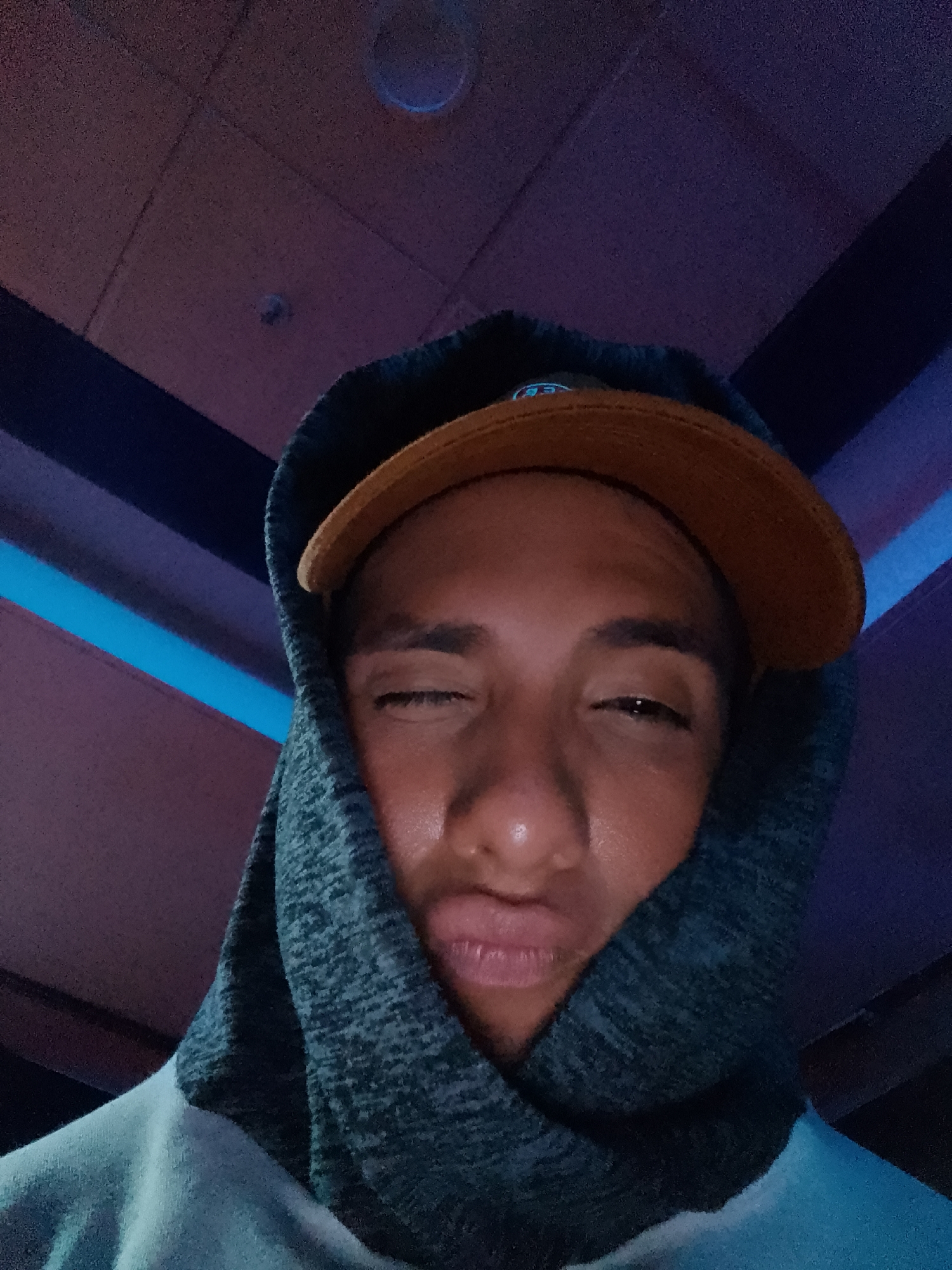 A close-up, low-angle shot features a man wearing a brown cap and a blue hooded sweatshirt. The hood is pulled up tightly around his face. The man's eyes are nearly closed, and he has a slight pucker to his lips. The background consists of ceiling tiles, some with blue lighting. The location in the city of Porlamar, Venezuela, cannot be determined from the image.