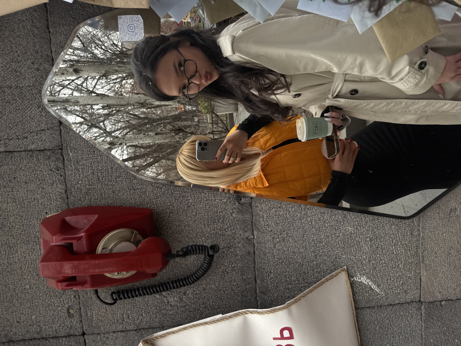 Two women are reflected in an ornate mirror. One woman wears a tan trench coat and glasses. The other has blonde hair and an orange vest, using her phone. A red rotary phone is mounted on the grey stone wall next to the mirror. A white tote bag with red lettering sits below the phone. Bare trees are visible in the background of the mirror's reflection.