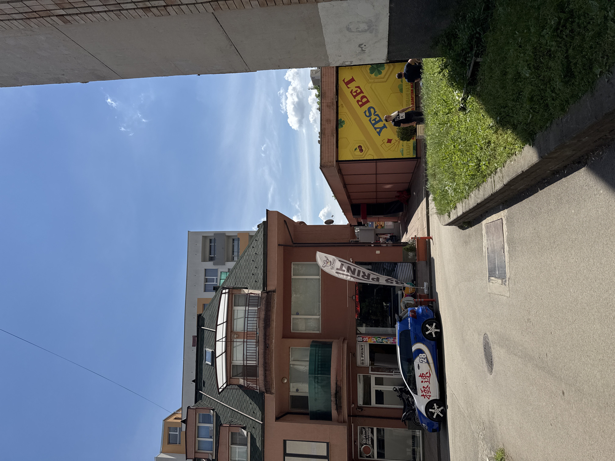 This image depicts a narrow street in Sofia, Bulgaria. A three-story building, partially obscured by an overhanging structure above, houses a business named "BS PRINT," as indicated by multiple signs on its facade and a waving flag. A blue and white car with stylized markings and the number "28" is parked along the street in front of the building. In the background, a yellow sign for "YES BET" is visible. Two people are standing near the betting sign. The sky is clear and blue with some scattered clouds. Grass and a concrete curb are along the right edge of the street.