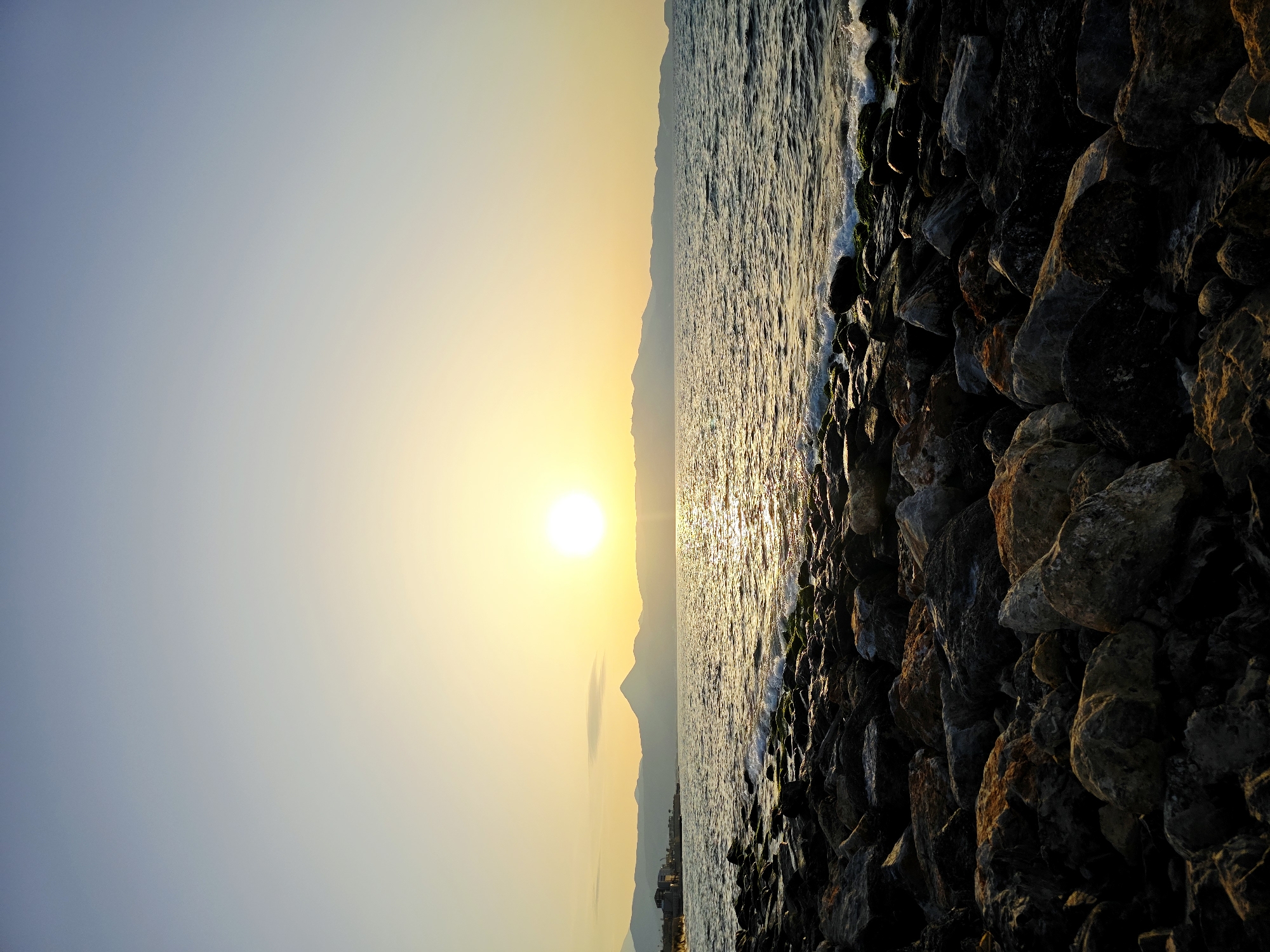 The image depicts a serene seascape at sunset in the city of Heraklion Municipal Unit, Greece. In the foreground, a rocky shore is covered with various-sized stones, some showing hints of moss. The sea, with gentle waves lapping against the rocks, stretches out towards the horizon. A faint line of coastal buildings and distant mountains can be seen on the left side of the frame. The sun, a bright orb, is positioned in the upper right quadrant of the sky, casting a golden glow and its reflection shimmers across the water. The sky transitions from a pale yellow near the horizon to a soft blue overhead, indicative of the twilight hour. There are no people or animals visible in the scene.
