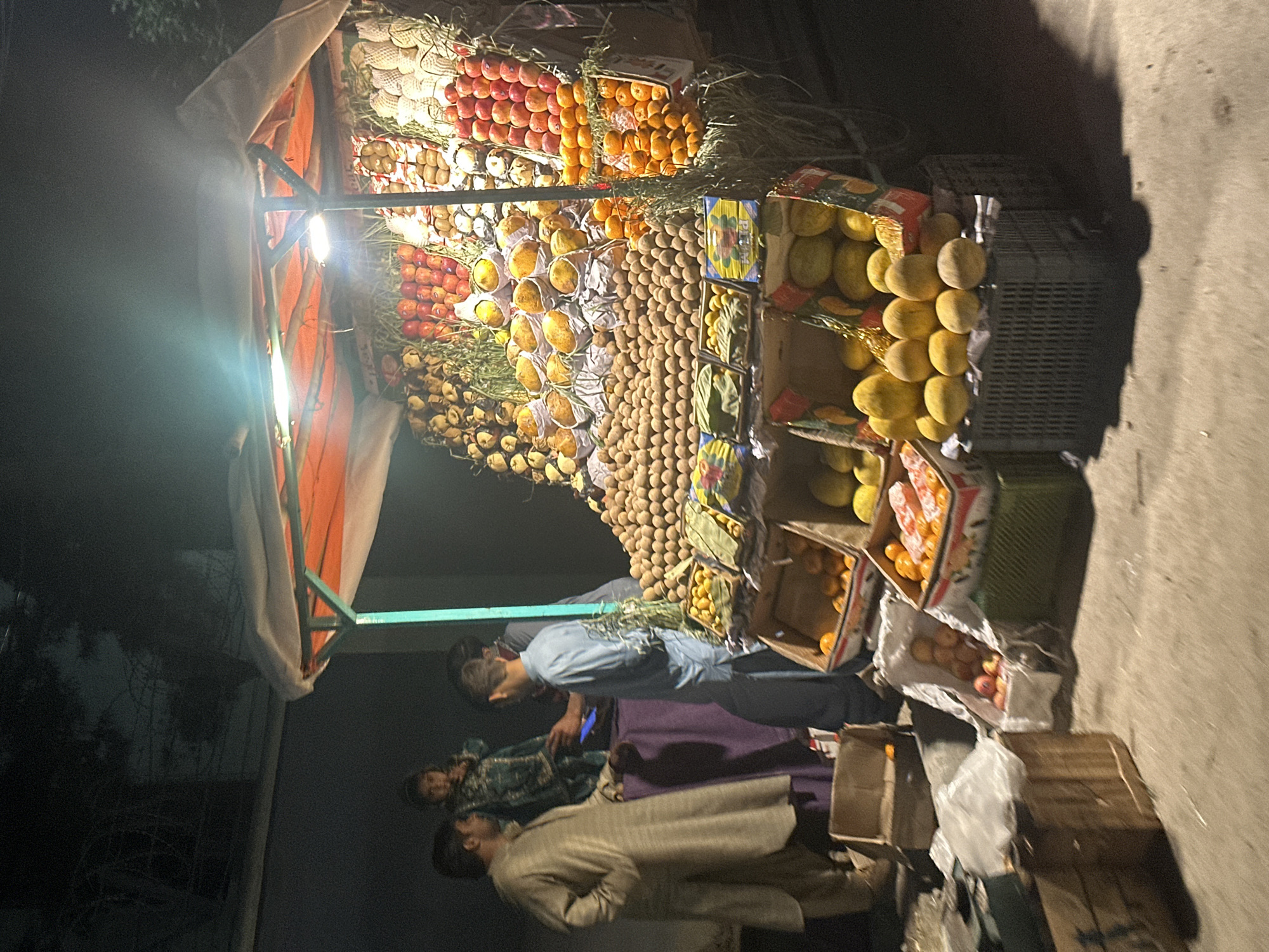 The image shows a fruit stand in Quetta City Tehsil, Pakistan. The stand is brightly lit and laden with a variety of fruits, including apples, oranges, mangoes, and longans, arranged in boxes and displays. Three people are present: two men appear to be customers, and one man is likely the vendor, standing behind the stall and interacting with the customers. The surrounding environment is dimly lit, suggesting it is evening or night. The stand is situated on a paved surface, likely a street or sidewalk. Additional crates and boxes are seen at the base of the stand.