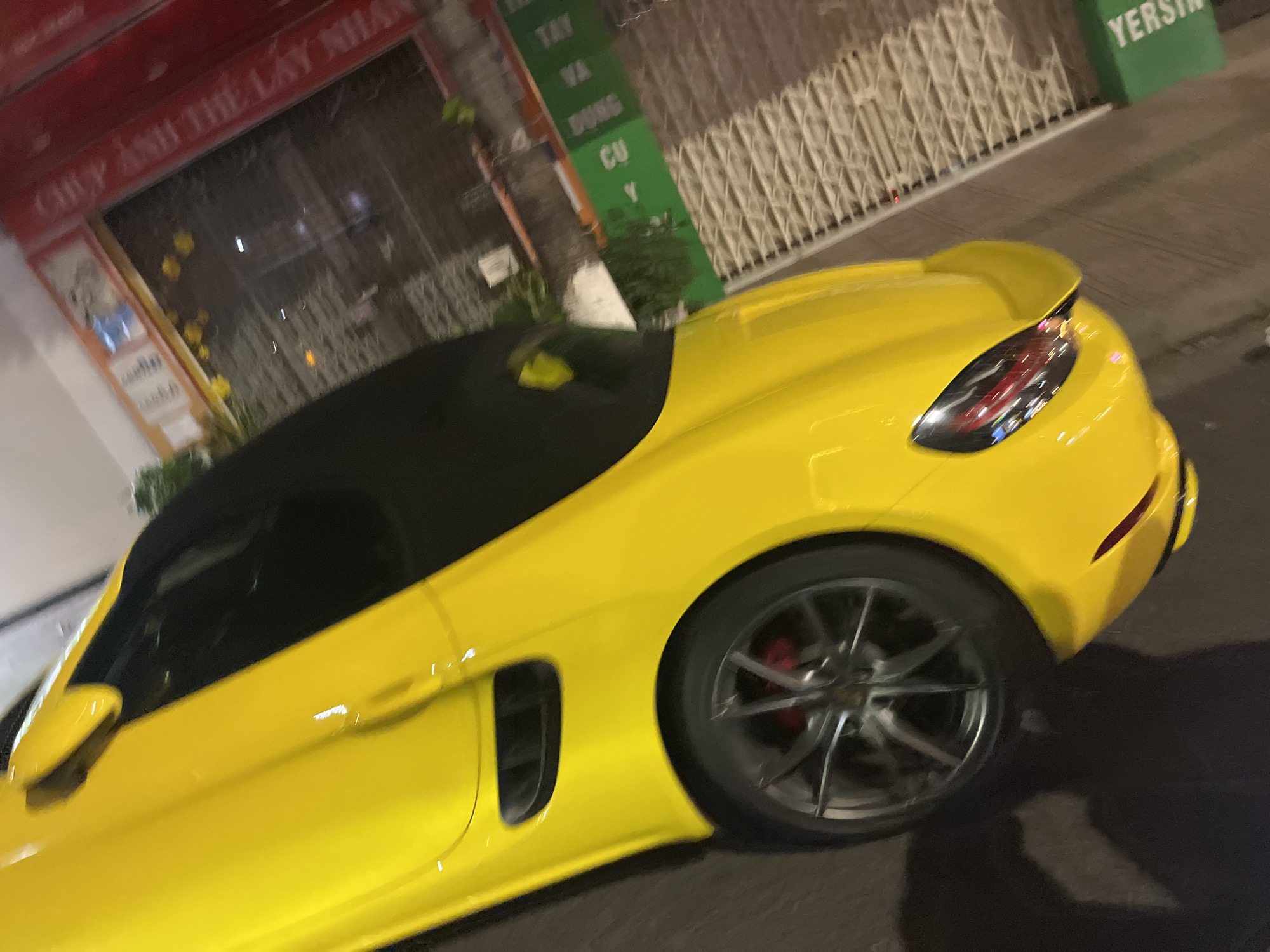 A yellow convertible car is parked on the side of a street in what appears to be an urban area. The car is angled towards the right side of the frame and has a black roof. The rear wheel and brake rotor are visible. The background features buildings with signs in Vietnamese, including a red sign on one building and a green sign on another. The general location seems to be in Nha Trang Ward, Vietnam. The street appears to be well-lit.
