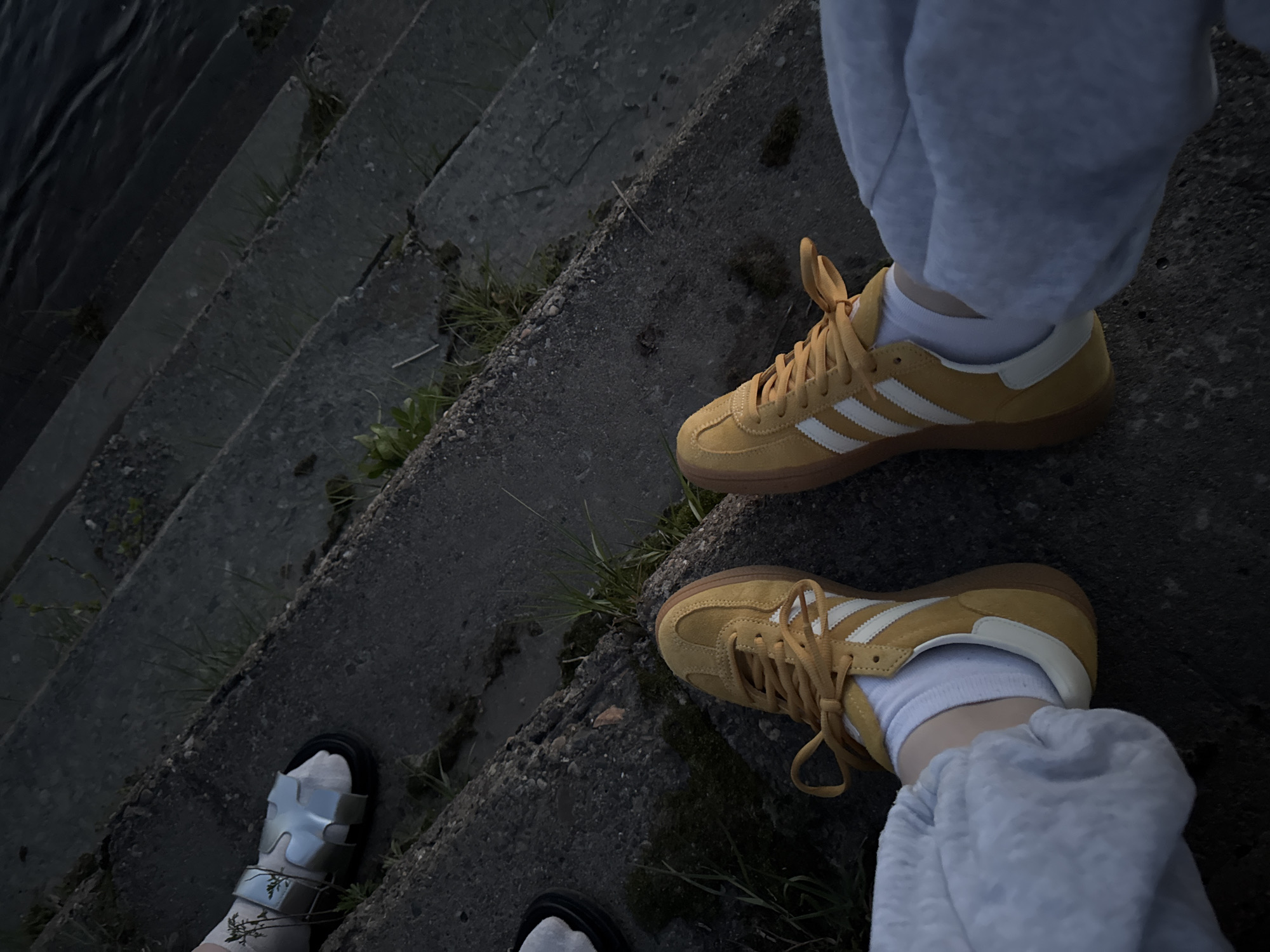 The image shows a view of concrete steps near water, with two pairs of feet visible. One pair is wearing yellow Adidas sneakers with white stripes, white socks, and grey sweatpants. The other pair is wearing silver sandals with white socks. The steps are weathered and have some patches of moss or vegetation growing on them. The water is visible on the left side of the frame.