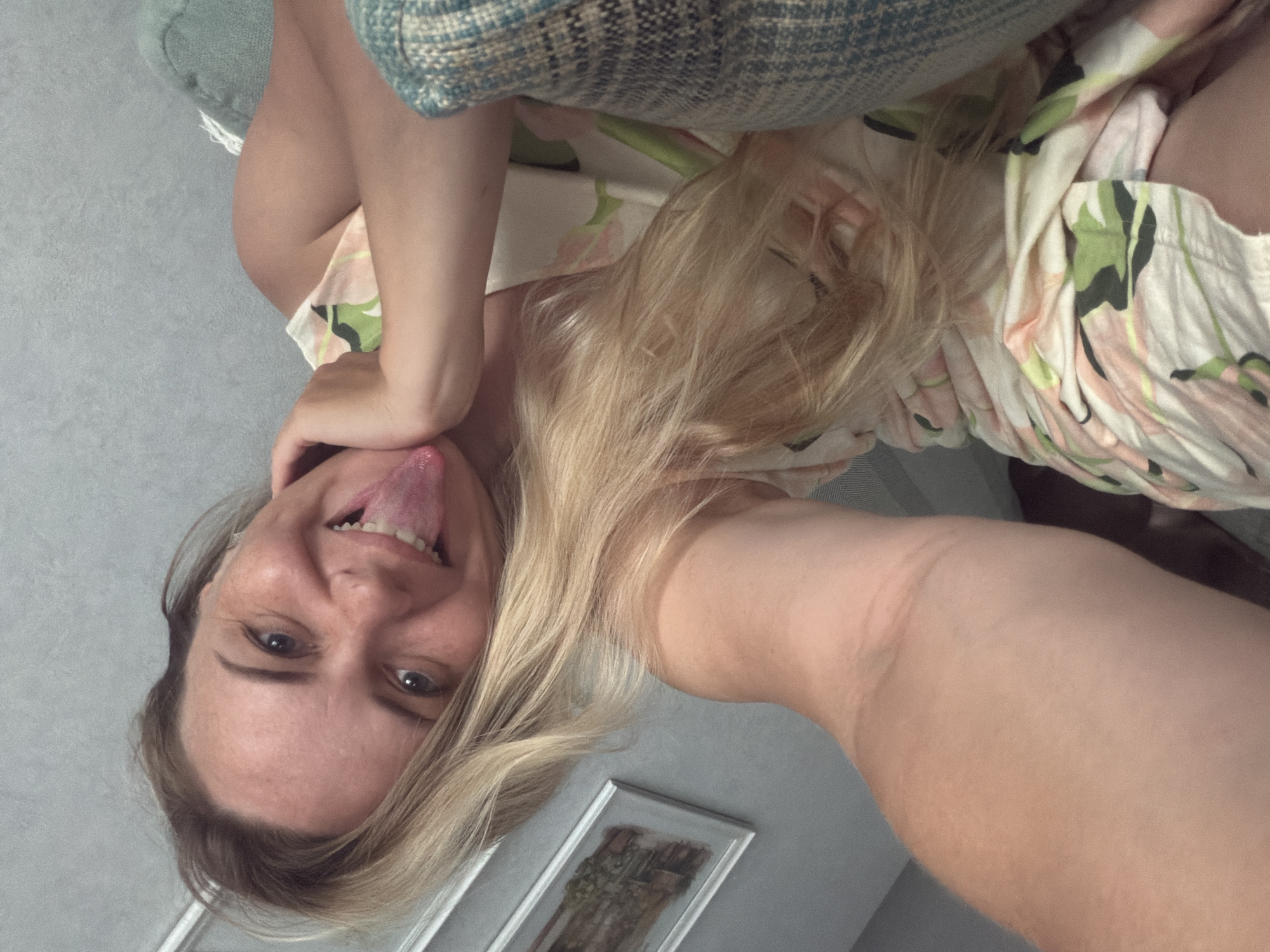 A woman with blonde hair and fair skin is seen in a close-up selfie, sticking out her tongue. She is wearing a light-colored dress with a floral print. The background appears to be an indoor setting, possibly a living room, with a textured wall and a glimpse of a painting on the wall. The image is taken from a low angle, with a portion of the woman's arm in the foreground. No specific location in Votkinsk, Russia, can be determined from the image.
