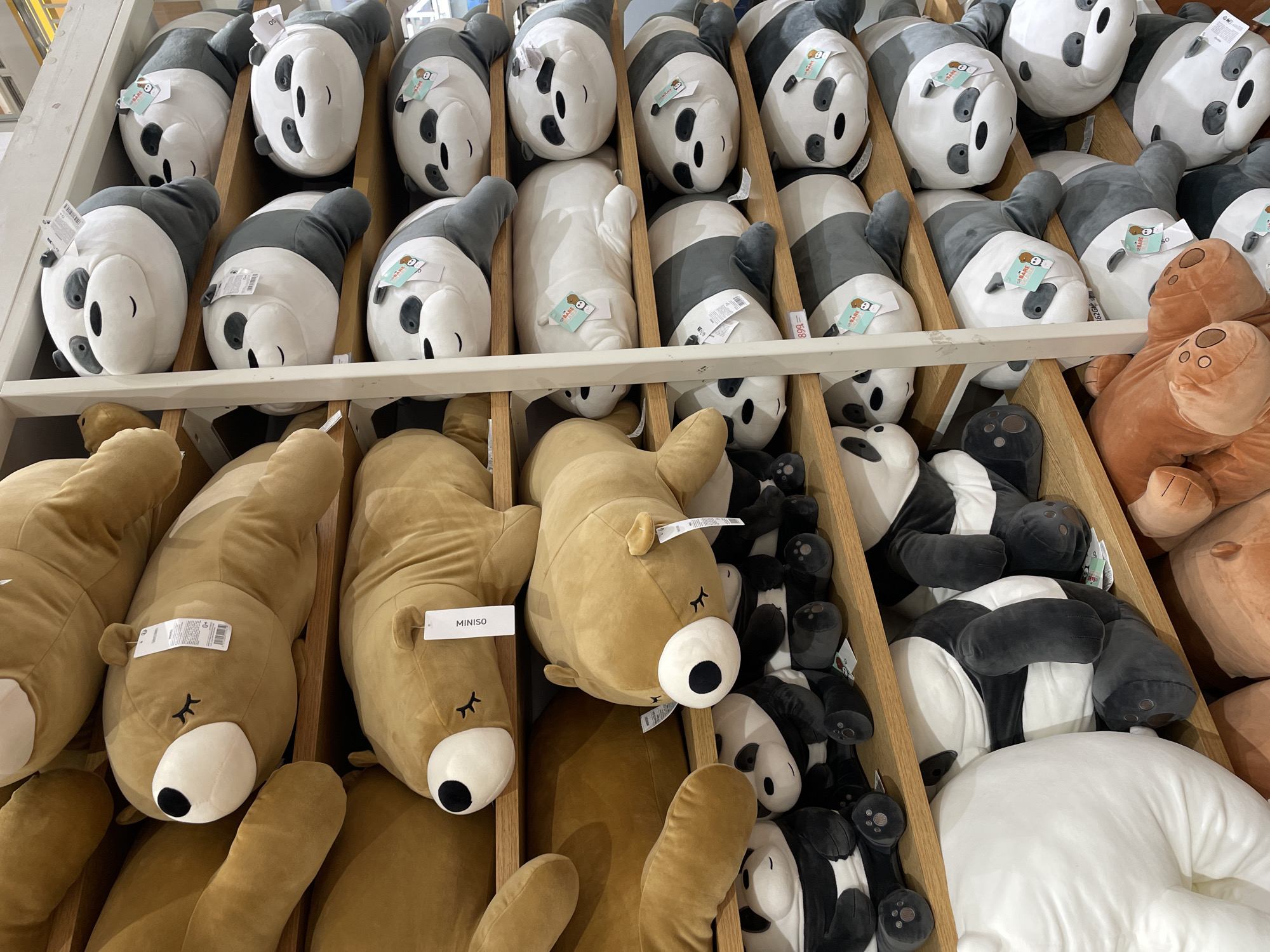 The image depicts a retail display of stuffed animals, specifically pandas and bears, arranged on wooden shelves. The stuffed animals are predominantly brown bears and black-and-white pandas, with various sizes and poses. Some have tags indicating they are from MINISO. The lighting suggests an indoor environment, likely a store. The scene is a close-up view of the merchandise, with the focus on the plush toys. There is no indication of the specific location within Davao City, Philippines; this information cannot be determined from the visual content provided.