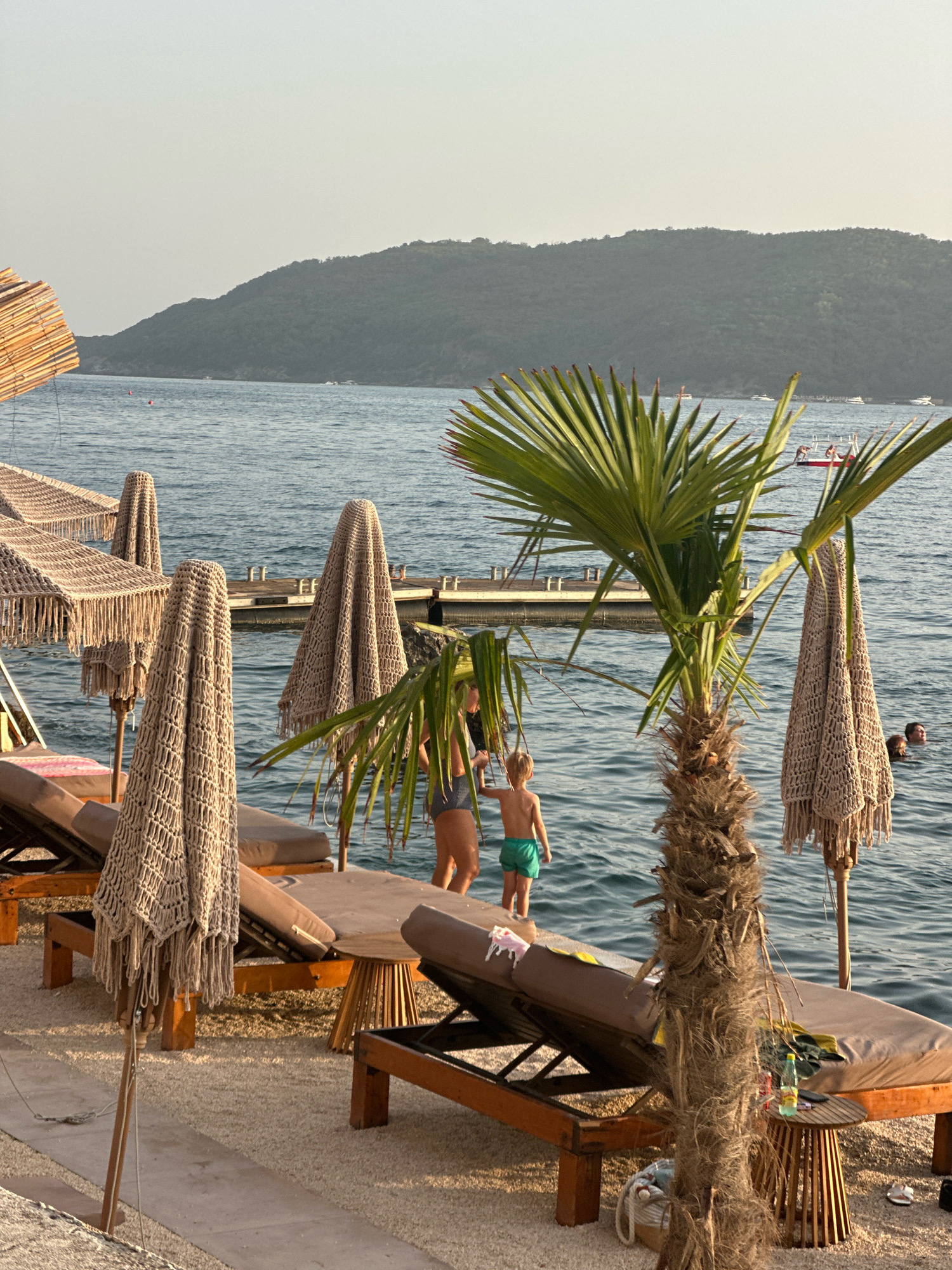 This image captures a serene beach scene in Budva, Montenegro, likely during the late afternoon given the soft, warm lighting. In the foreground, several empty sun loungers with brown cushions are neatly arranged on a gravelly surface, hinting at a well-maintained beach resort. Decorative umbrellas with macrame detailing and fringe are set up, adding a bohemian touch to the scene. A young boy wearing green swim trunks stands near the water's edge, his back to the camera, looking out at the sea. Further out in the water, an adult woman with dark hair is visible, also facing away from the viewer, and the heads of two other individuals are barely breaking the surface of the water, suggesting they are swimming or relaxing.
The middle ground is dominated by the calm, blue sea. A wooden pier with metal railings extends into the water, and a small red boat with people on it is docked near the pier. In the distance, a range of lush, green hills forms a picturesque backdrop, their slopes covered in vegetation. The sky is a pale, hazy blue, characteristic of a warm, clear day. The overall impression is one of tranquility and relaxation, with elements of both natural beauty and curated comfort.