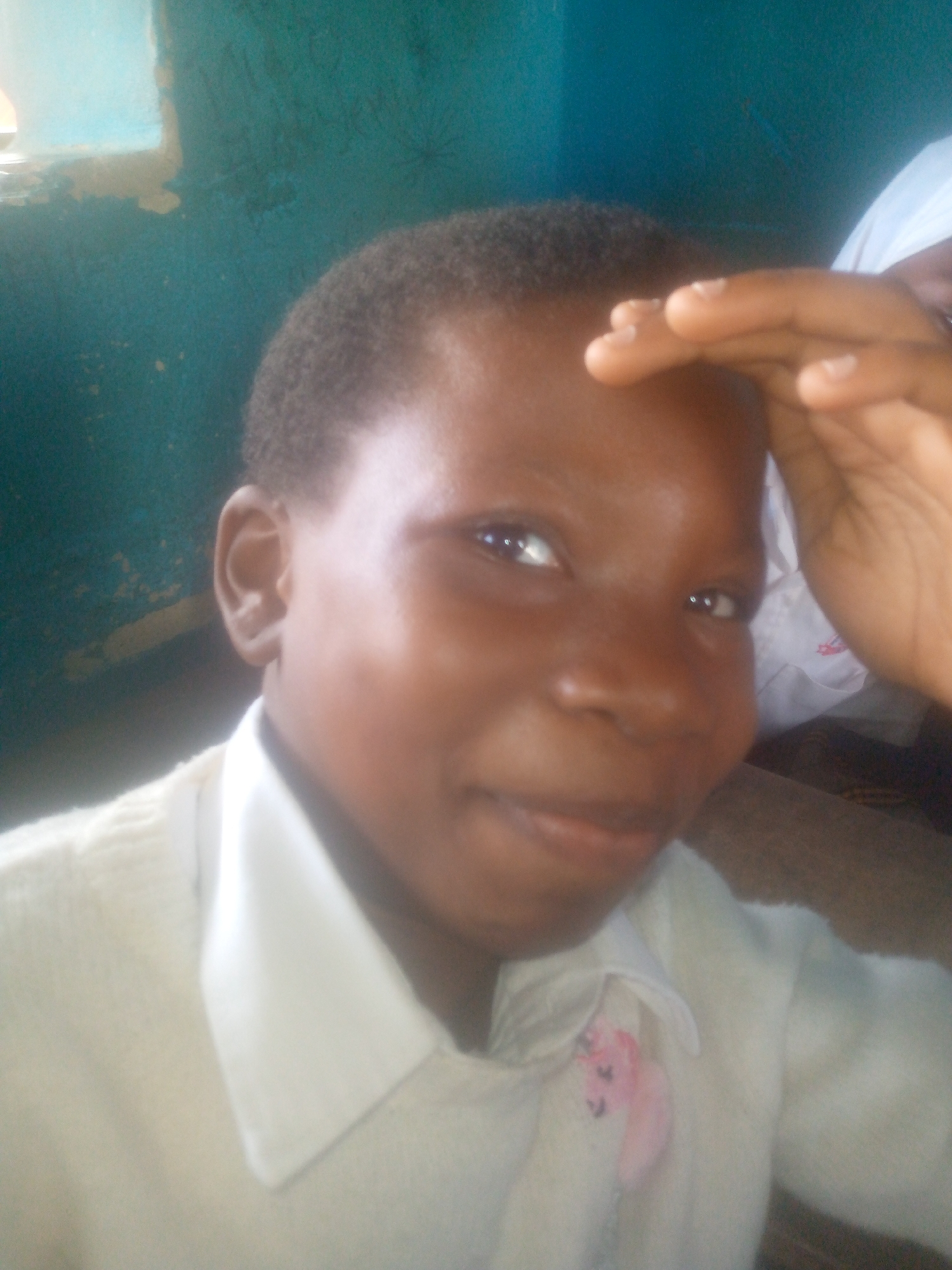 A young, dark-skinned person is facing the camera with their right hand raised towards their forehead. The person has short, dark hair. They are wearing a light-colored collared shirt with a pink stain near the collar. The background is a teal-colored wall, with part of a window in the upper left corner. Another person, wearing a white shirt, is partially visible to the right of the subject. The specific location in Ikorodu, Nigeria, is not discernable from the image.