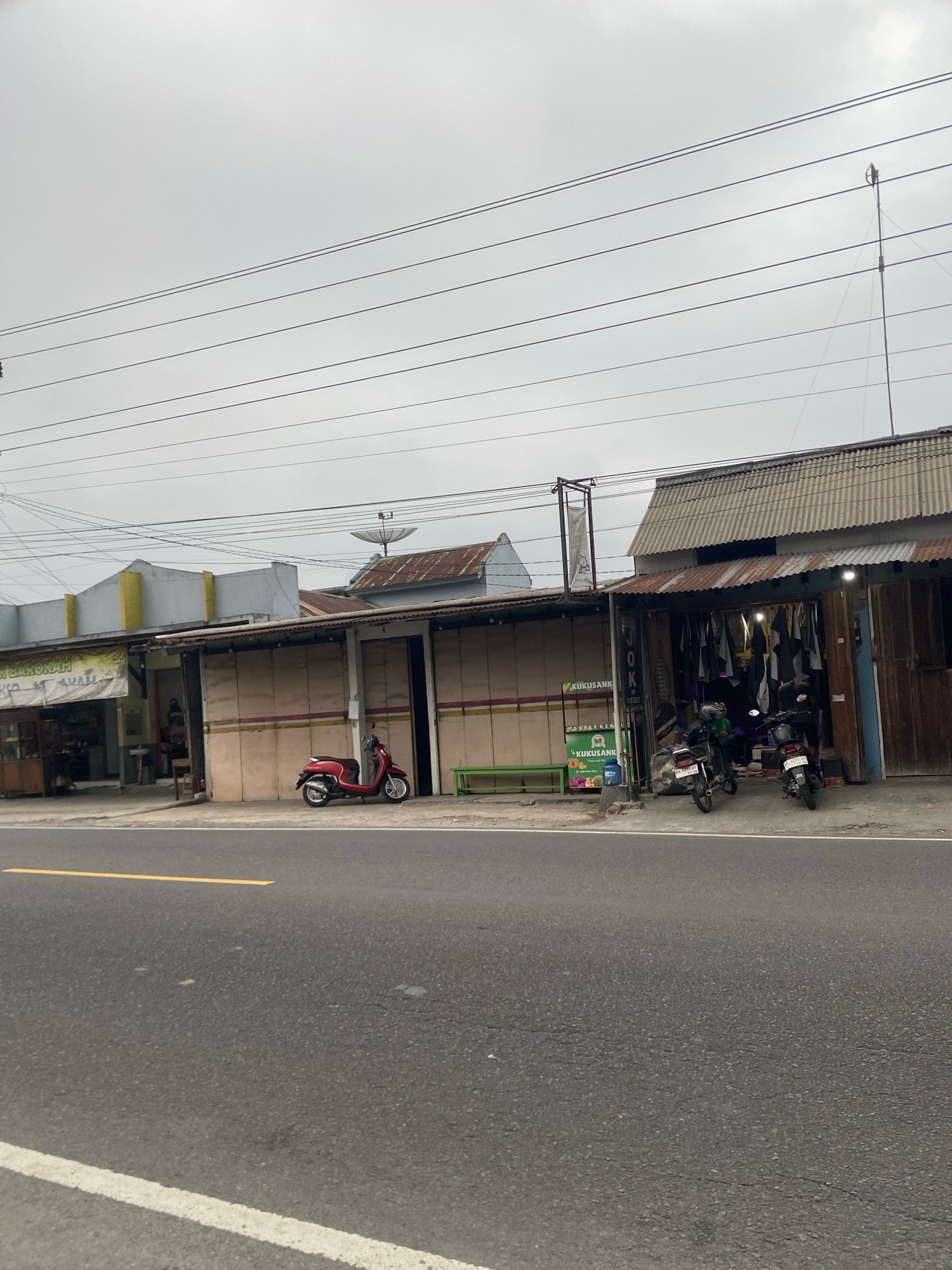 The image depicts a street scene in Karangluhur, Indonesia, under an overcast sky. In the foreground, a two-lane asphalt road with a yellow and white line marking is visible. The middle ground features a row of shops with corrugated metal roofs and light-colored walls, some with shuttered entrances. A red scooter is parked in front of one of the shops. To the right, another shop displays hanging garments, with two motorcycles parked outside. A satellite dish is mounted on a rooftop further in the background. Overhead, multiple power lines stretch across the frame. There are no people clearly visible in the image.