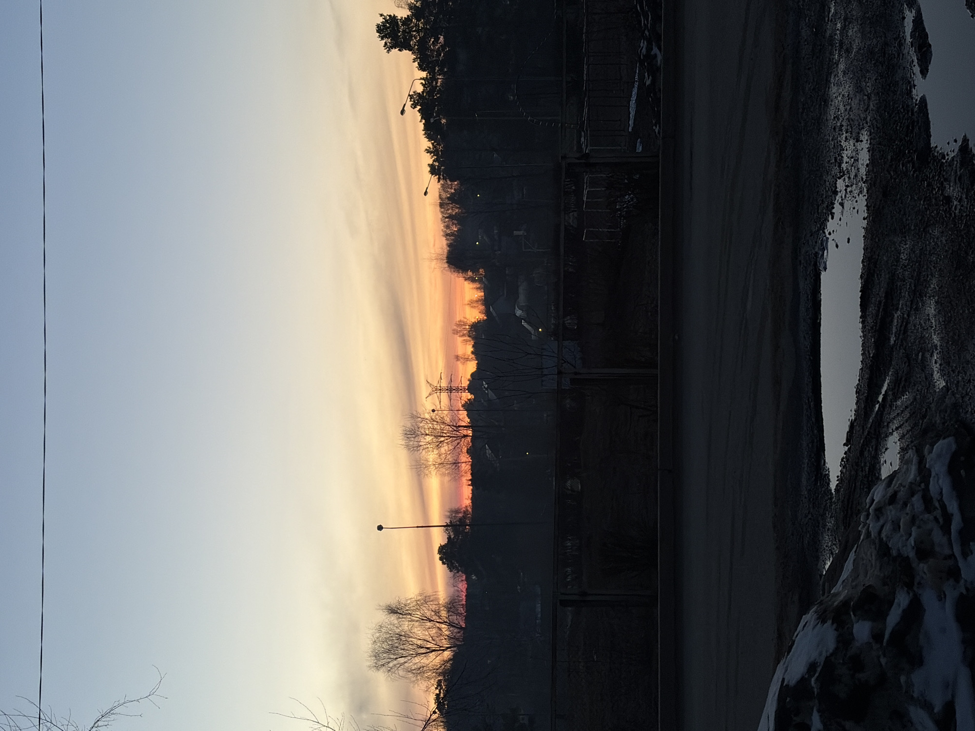The image depicts a sunset view from what appears to be a building in Chaikovsky, Russia. The sky transitions from light blue to orange and pink hues near the horizon, with cloud formations. Silhouetted trees, buildings, and a power transmission tower are visible against the colorful sky. A road runs horizontally across the middle of the image, with a sidewalk or edge of the road covered with snow and puddles. Lampposts are present on the roadside. A metal fence or railing is visible in front of the viewpoint.