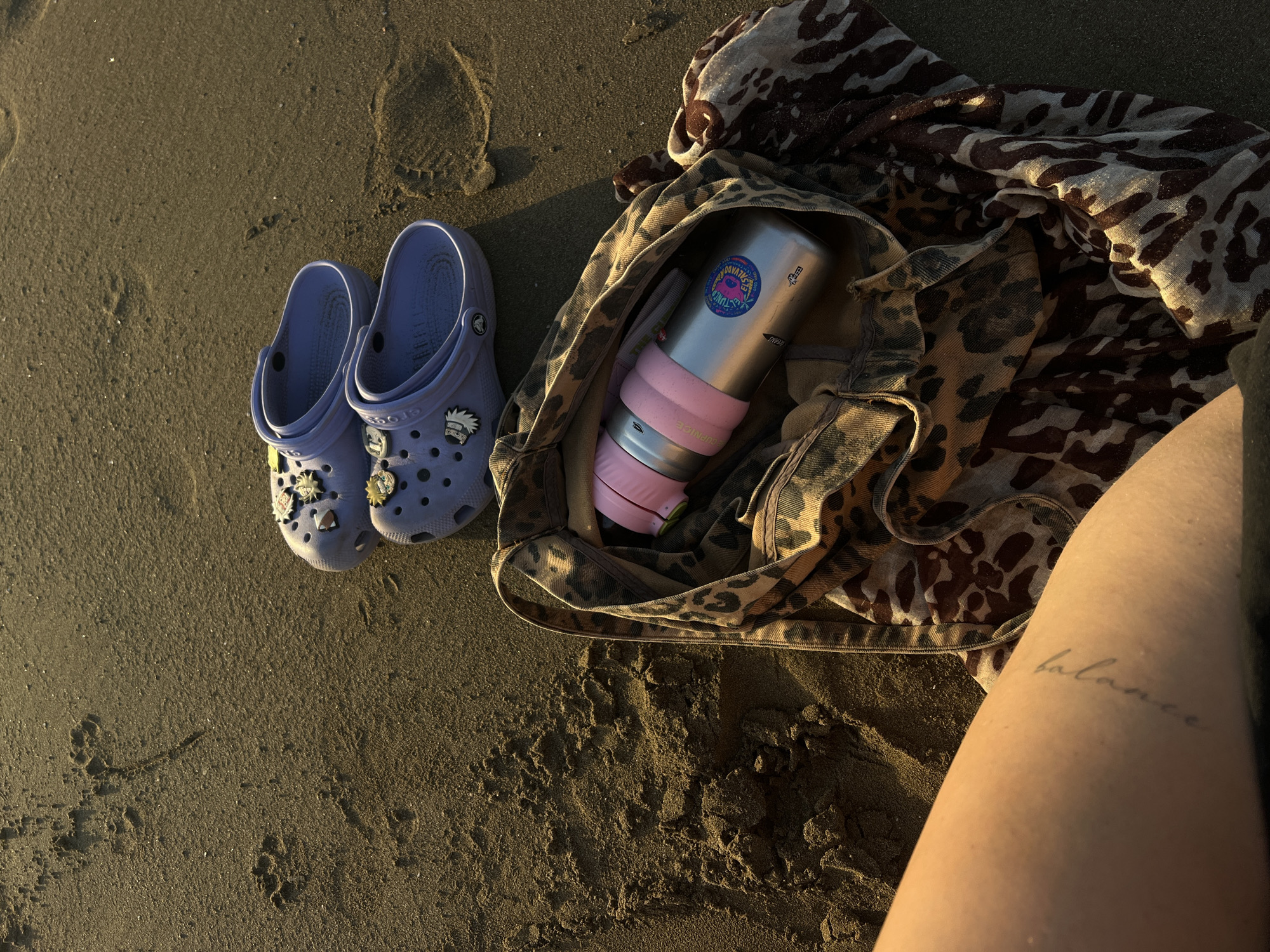 This is a close-up, overhead shot of a beach scene in San Juan del Sur, Nicaragua, with a person's lower left leg and arm visible in the foreground. The skin on the arm has a tattoo of the word "balance" in script.
Scattered on the damp, sandy beach are a pair of light purple Crocs with various charms, a leopard-print tote bag, and a leopard-print scarf. Inside the tote bag, a stainless steel water bottle with a pink grip is visible. The water bottle has a sticker on it with the text "El Salvador". The sand has visible footprints and texture from being near the water. The lighting suggests it is either early morning or late afternoon.