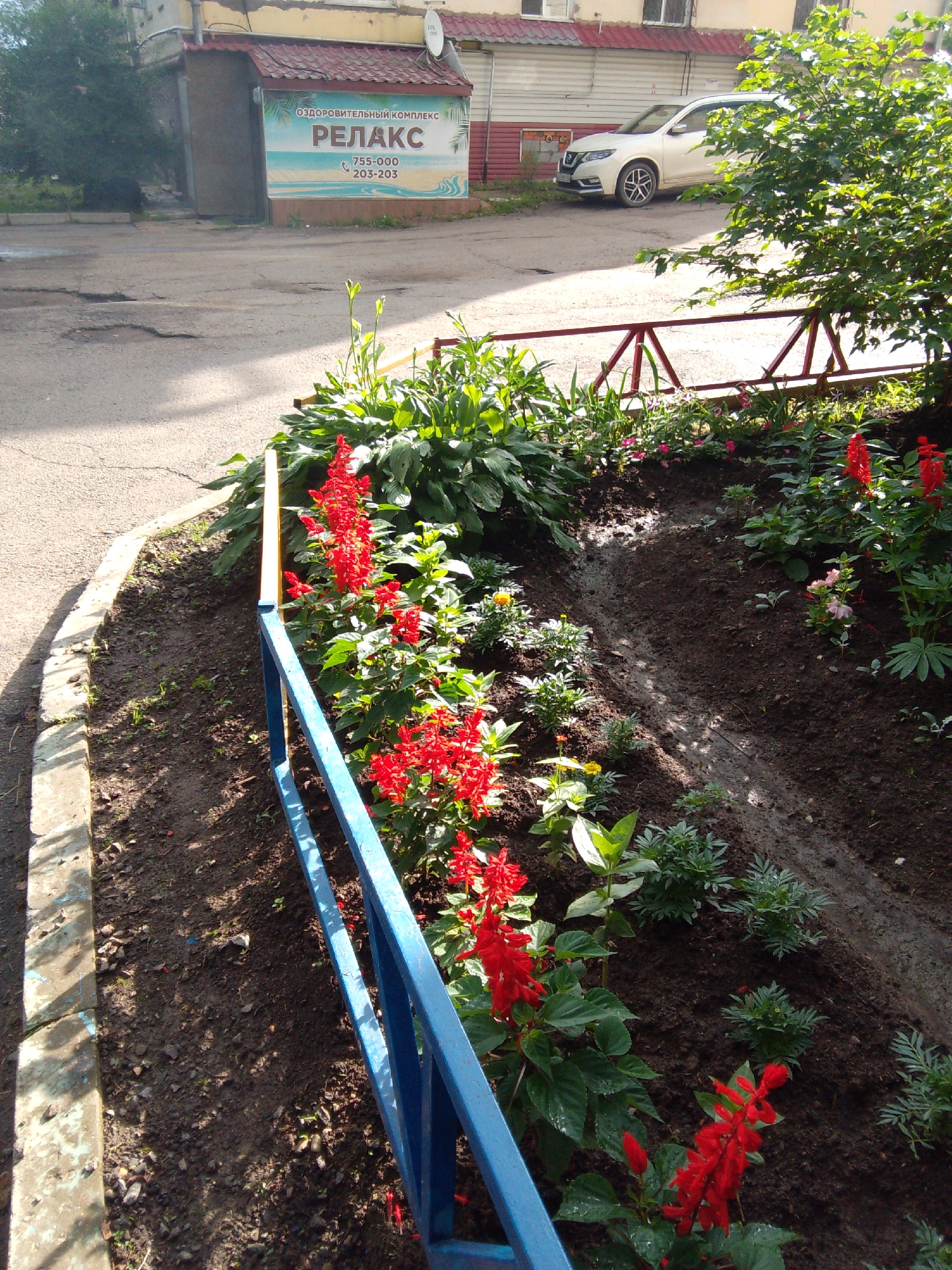 The image shows a residential building with a sign for "RELAX" health complex in Khabarovsk, Russia. In front of the building, there is a flower bed with red salvia and other green plants, bordered by a blue and yellow fence. A white SUV is parked on the asphalt road in front of the building. The ground is wet, suggesting recent rain.