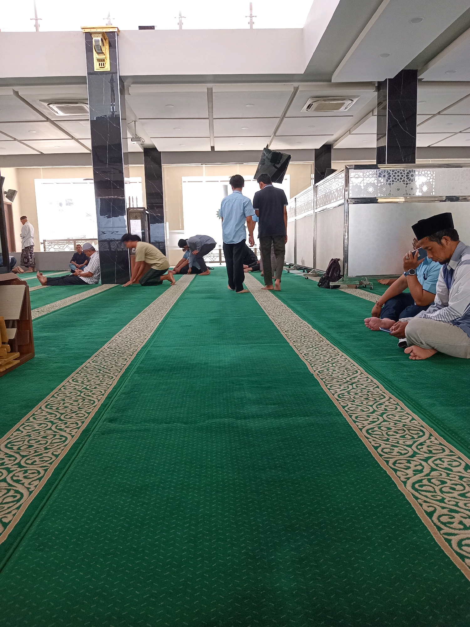 The image depicts the interior of a mosque in Randuagung, Indonesia. The floor is covered with a green carpet with intricate beige patterns. Several men are present, some standing, others sitting or kneeling on the carpet. Two men in the foreground on the right are sitting and looking at their phones. One man in the center is walking away from the camera, while others are dispersed throughout the mosque, engaged in prayer or conversation. The mosque's architecture features high ceilings with modern lighting and air conditioning units. Dark marble columns support the ceiling, and a decorative wall with Islamic geometric patterns separates sections of the interior. The overall atmosphere suggests a place of worship during a time of prayer or congregation.