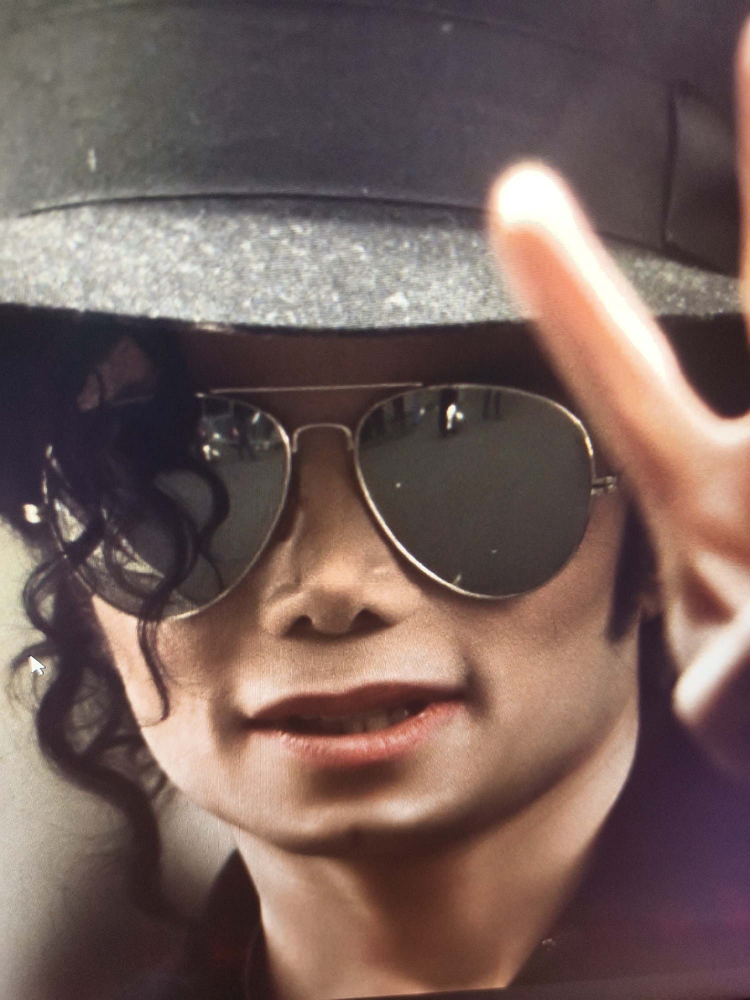 A close-up portrait features Michael Jackson, identifiable by his distinctive features, wearing a grey fedora-style hat and aviator sunglasses. His face is centered, showing from his hat line down to his upper chest. He has dark, curly hair visible around the left side of his face. His mouth is slightly open, revealing part of his teeth, and he displays a subtle, relaxed expression. The reflective lenses of his sunglasses obscure his eyes, reflecting blurred elements from the surroundings.
On the right side of the frame, a hand is partially visible, with two fingers raised, possibly forming a peace sign or a casual gesture. These fingers are brightly illuminated, creating a soft glow around their edges. The background is out of focus, appearing dark and indistinct, suggesting an indoor setting or controlled environment.
The image itself appears to be a photograph of a digital display, evidenced by a visible pixel grid pattern and a white mouse cursor on the left side of Michael Jackson's face. There is no visible text within the image. No specific time of day or weather can be determined, and the scene offers no visual cues for its location in Svilengrad, Bulgaria, beyond the provided context.