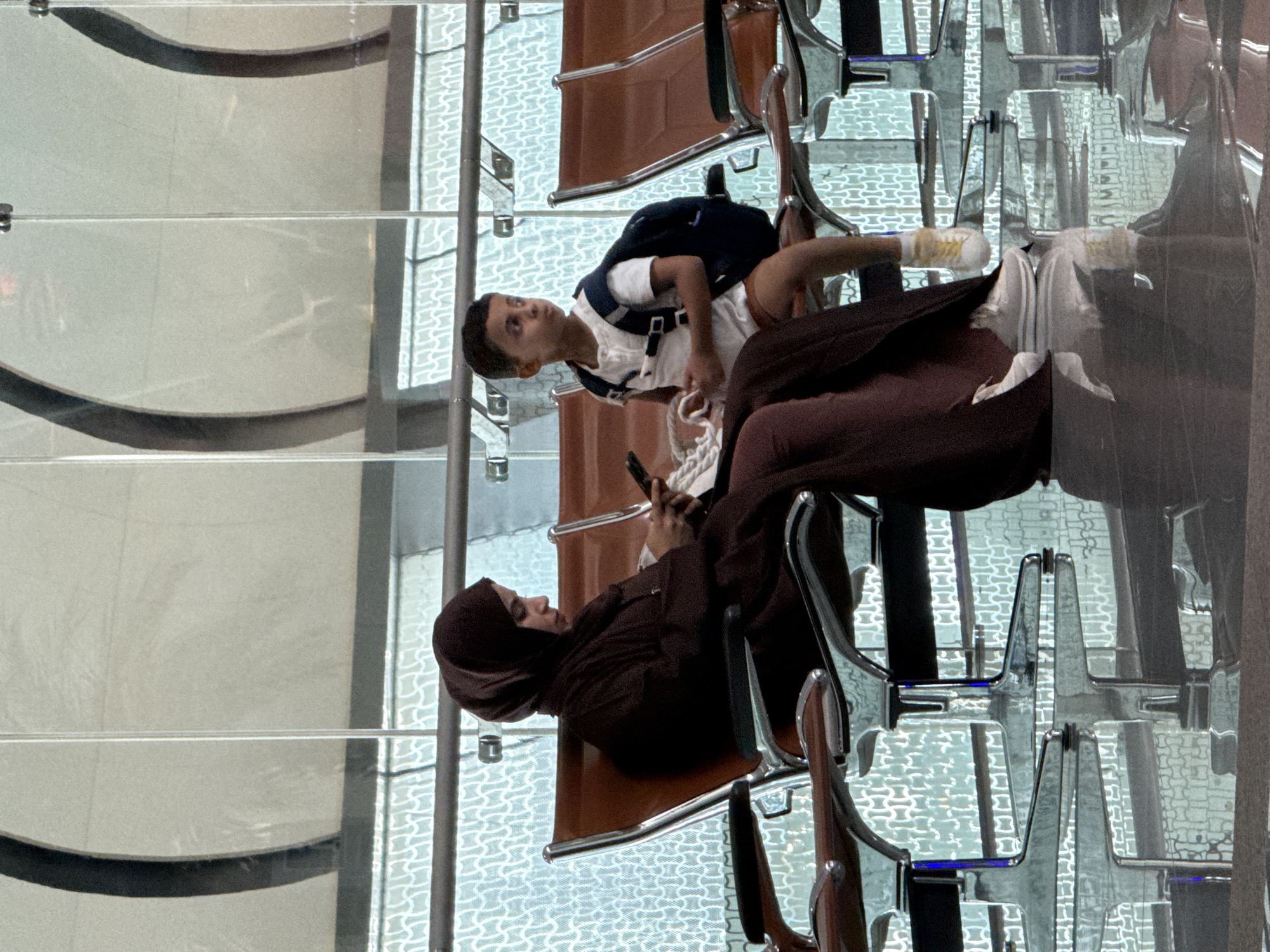 In Seeb Province, Oman, at what appears to be an airport lounge, a woman wearing a dark maroon abaya and hijab is seated, looking down at a smartphone in her hands. Beside her, a young boy in a white t-shirt and brown shorts is seated, wearing a dark backpack. He is looking away from the camera. Both are seated on brown airport chairs with chrome legs. The floor is highly reflective, mirroring the chairs and the patterned glass wall behind them. The environment suggests a public waiting area, likely within an airport due to the seating arrangement and the expansive glass structure.