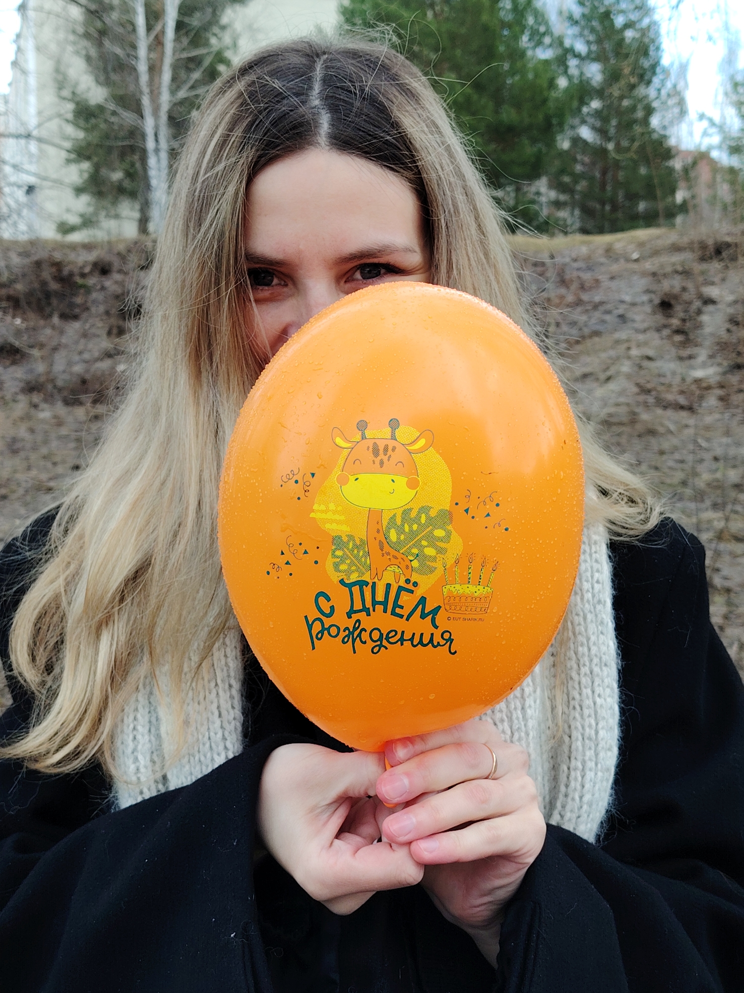 A fair-skinned woman with long, blonde, and partially wet hair holds an orange balloon in front of her face, partially obscuring it. The balloon has a cartoon giraffe printed on it, along with a cake and the inscription "С Днем Рождения" in Cyrillic letters. The woman is wearing a black coat and a white knitted scarf. Her eyes are visible above the balloon, and a wedding ring is on her finger. The background shows a blurred cityscape, including bare trees and a building. The image was taken in the city of Chaikovsky, Russia.