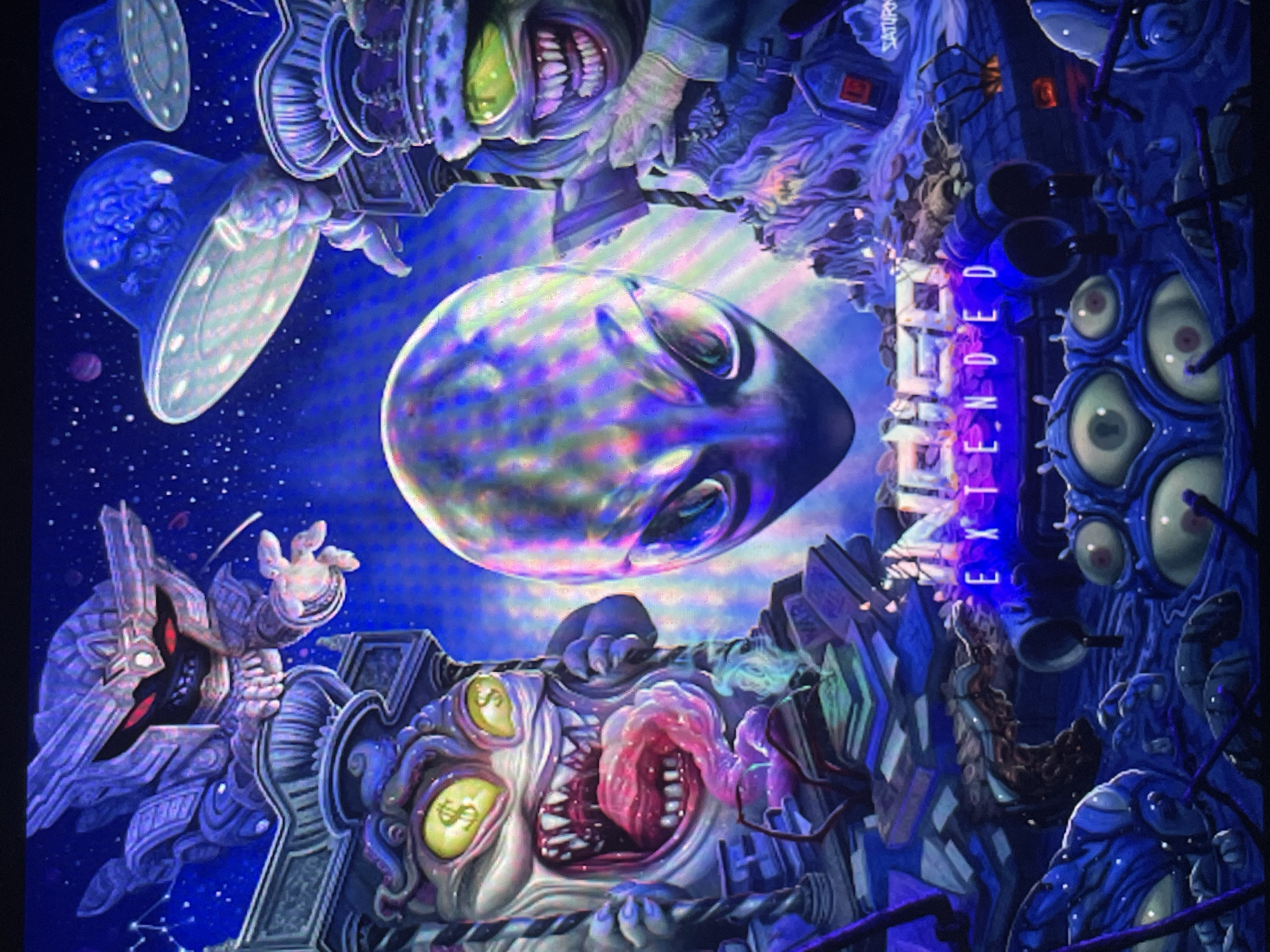 This intricate and vibrant artwork depicts a surreal, cosmic scene dominated by monstrous and alien forms. In the center, a large, iridescent orb or alien head with closed eyes glows against a deep blue, star-filled cosmic background.
To the top left, two transparent, bell-shaped UFOs float, each encasing a visible human brain, with a small planet in the distance. Above the central orb, an inverted, grotesque figure with green skin, sharp teeth, and a space-suit-like attire reaches out, with the word "SATURN" faintly visible above it.
On the bottom left, an angular, robotic figure with glowing red eyes and complex armor extends a hand. Below the central orb, a large, fleshy, multi-eyed creature dominates the foreground. Two of its yellow eyes feature dollar signs, and its wide-open mouth reveals a pink, tentacled tongue and sharp teeth.
To the right, a vertical, sprawling city-like structure appears, built from organic and mechanical elements, adorned with numerous bulbous eyes. A blue neon sign glows with the word "EXTENDED." Several small, dark, spider-like creatures are seen on these structures. The overall scene is chaotic and imaginative, blending elements of science fiction, horror, and fantasy, conveying an unsettling and bizarre atmosphere.