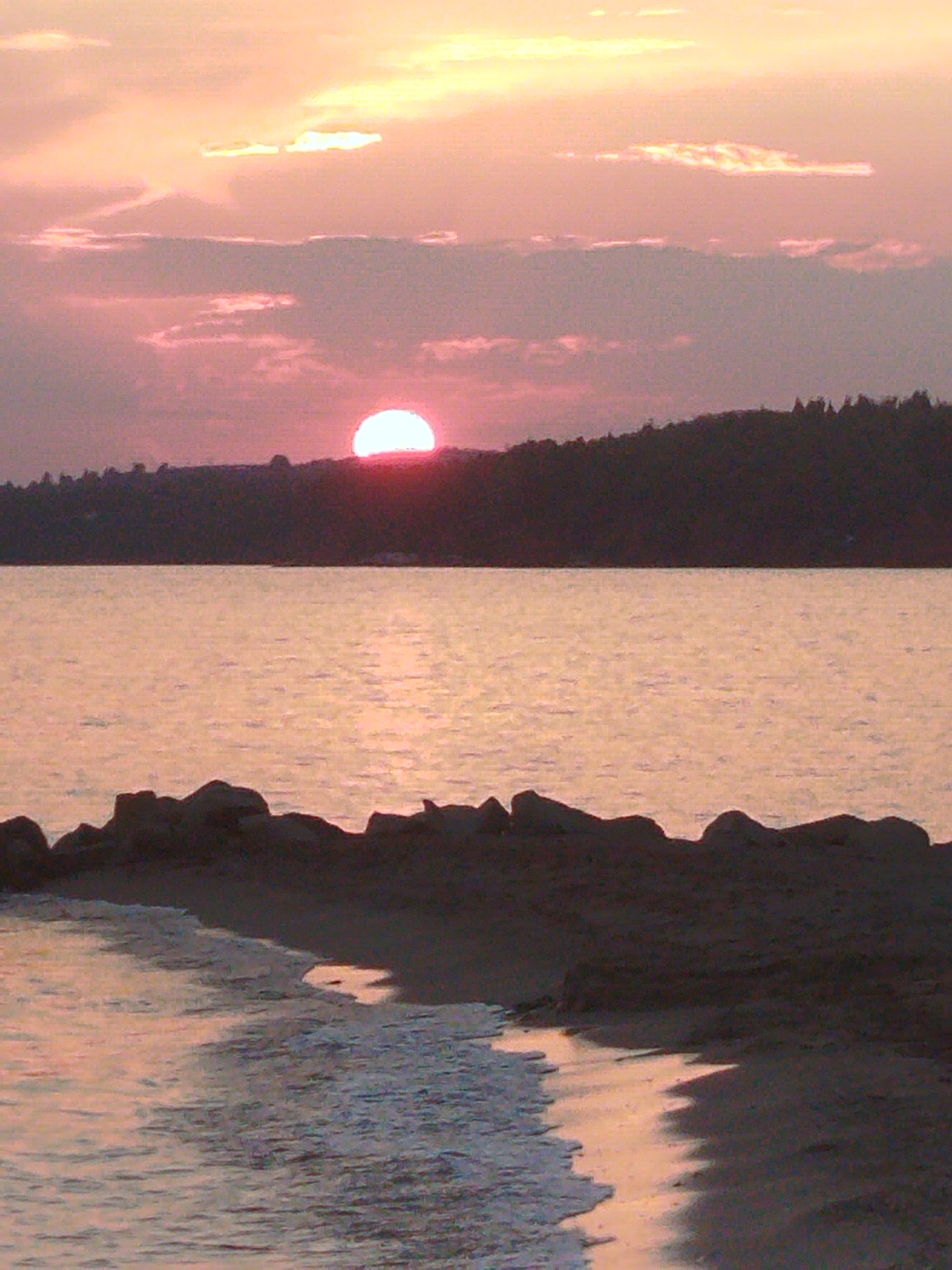 The image depicts a sunset over the sea in Sithonia Municipal Unit, Greece. The sun, partially obscured by clouds, casts a warm, pinkish glow across the sky and the water. The horizon is marked by a dark, silhouetted treeline on a hill. In the foreground, the gentle waves of the sea lap against a rocky shore. The overall atmosphere is serene and tranquil, capturing the natural beauty of the Greek coast at dusk.