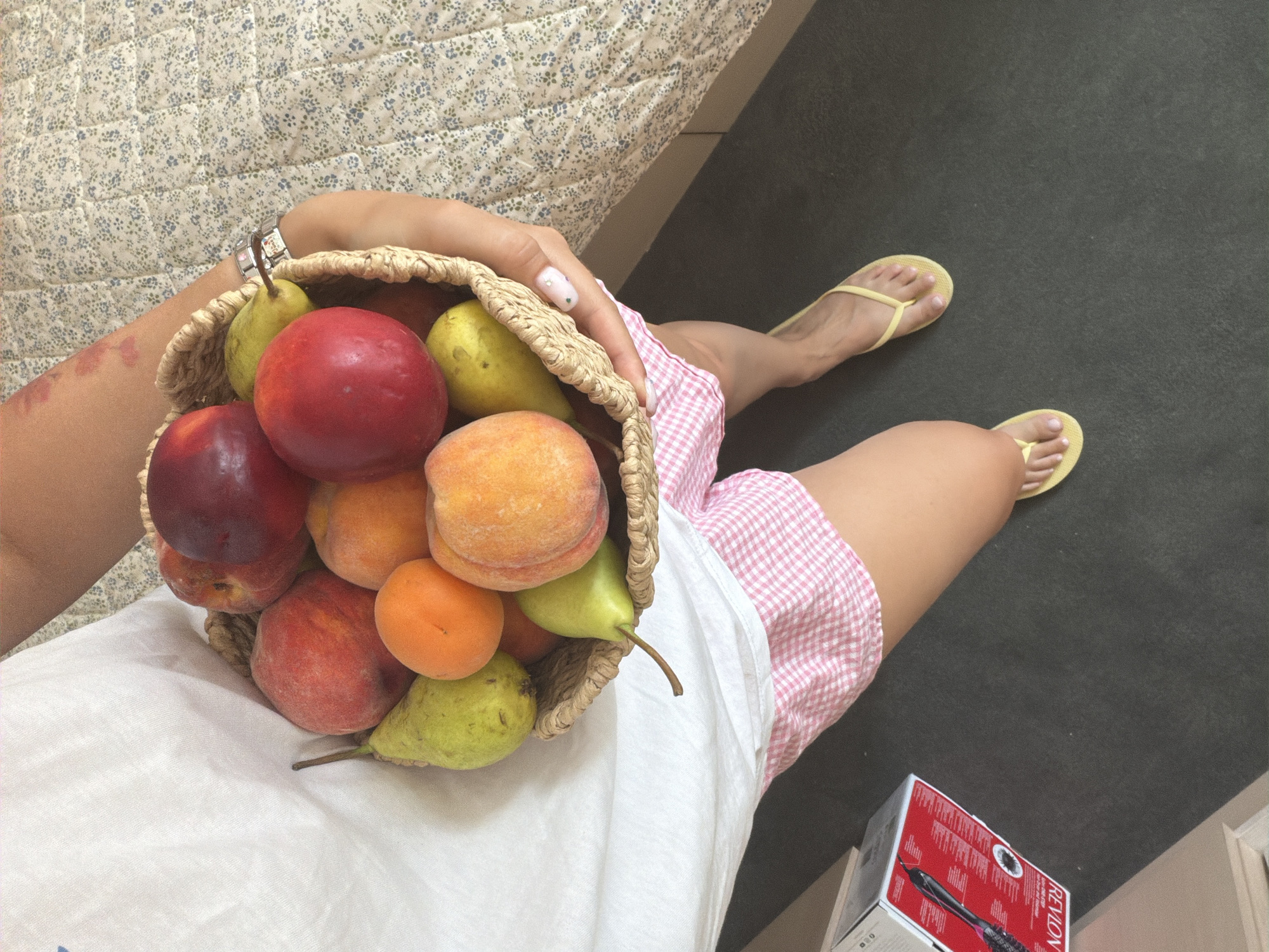 A person is holding a woven basket filled with assorted fruits, including peaches, pears, and apricots. They are wearing a white t-shirt and pink checkered shorts, along with yellow flip-flops. The background shows a bed with a floral patterned sheet and a dark grey carpet. A Revlon hair product box is visible on the floor to the left. The scene does not contain any specific indicators of its location within Sofia, Bulgaria.