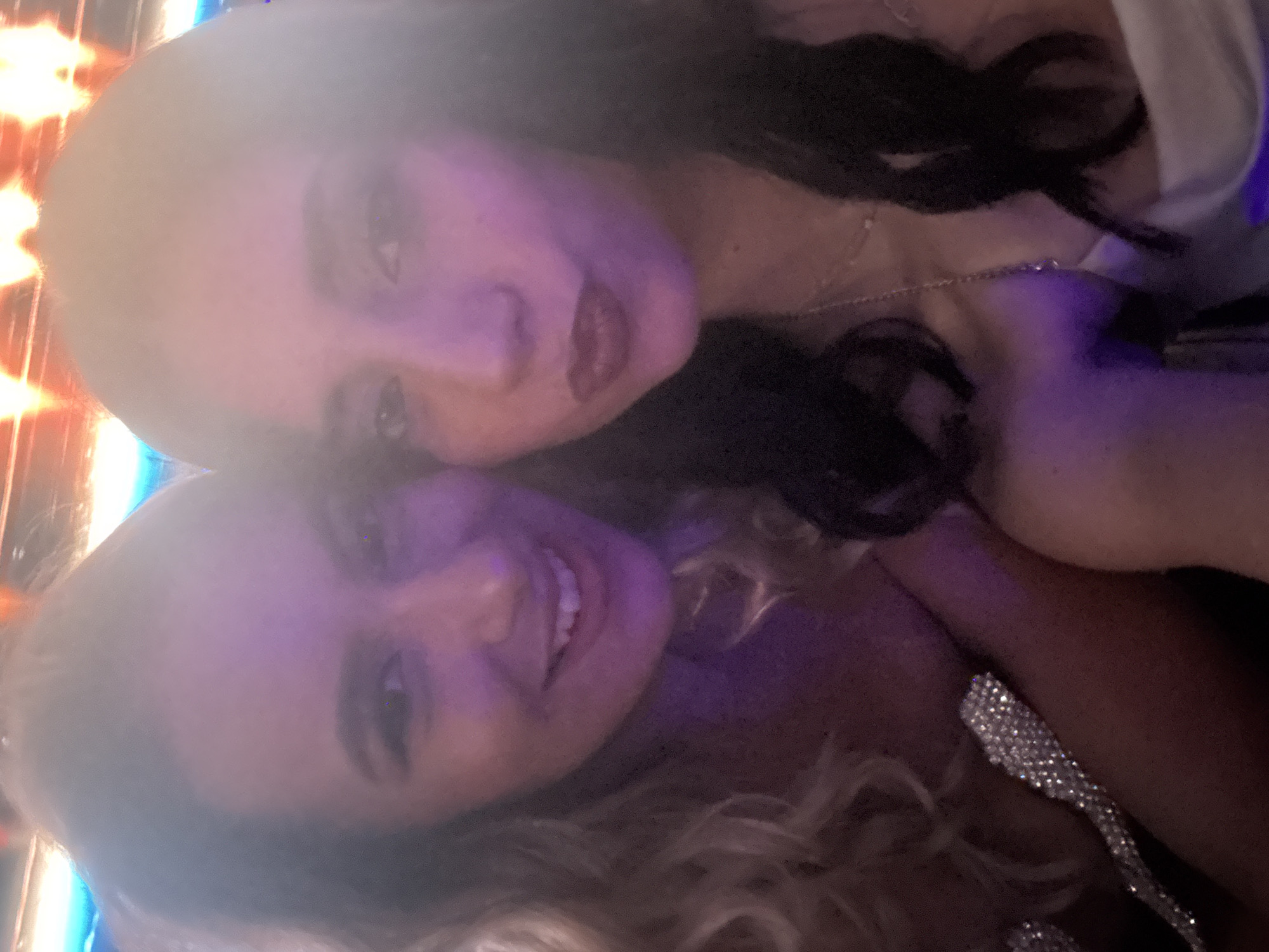 Two young women are taking a selfie indoors, likely at an event or nightclub given the lighting. The woman on the left has blonde, curly hair and is smiling. She is wearing a dress with sparkly embellishments on the strap. The woman on the right has dark, wavy hair and is wearing dark lipstick. She is also smiling. The background is blurred with colorful lights, suggesting a vibrant setting. The overall impression is one of a casual, fun social gathering. No specific location in Sofia, Bulgaria can be determined from the image.