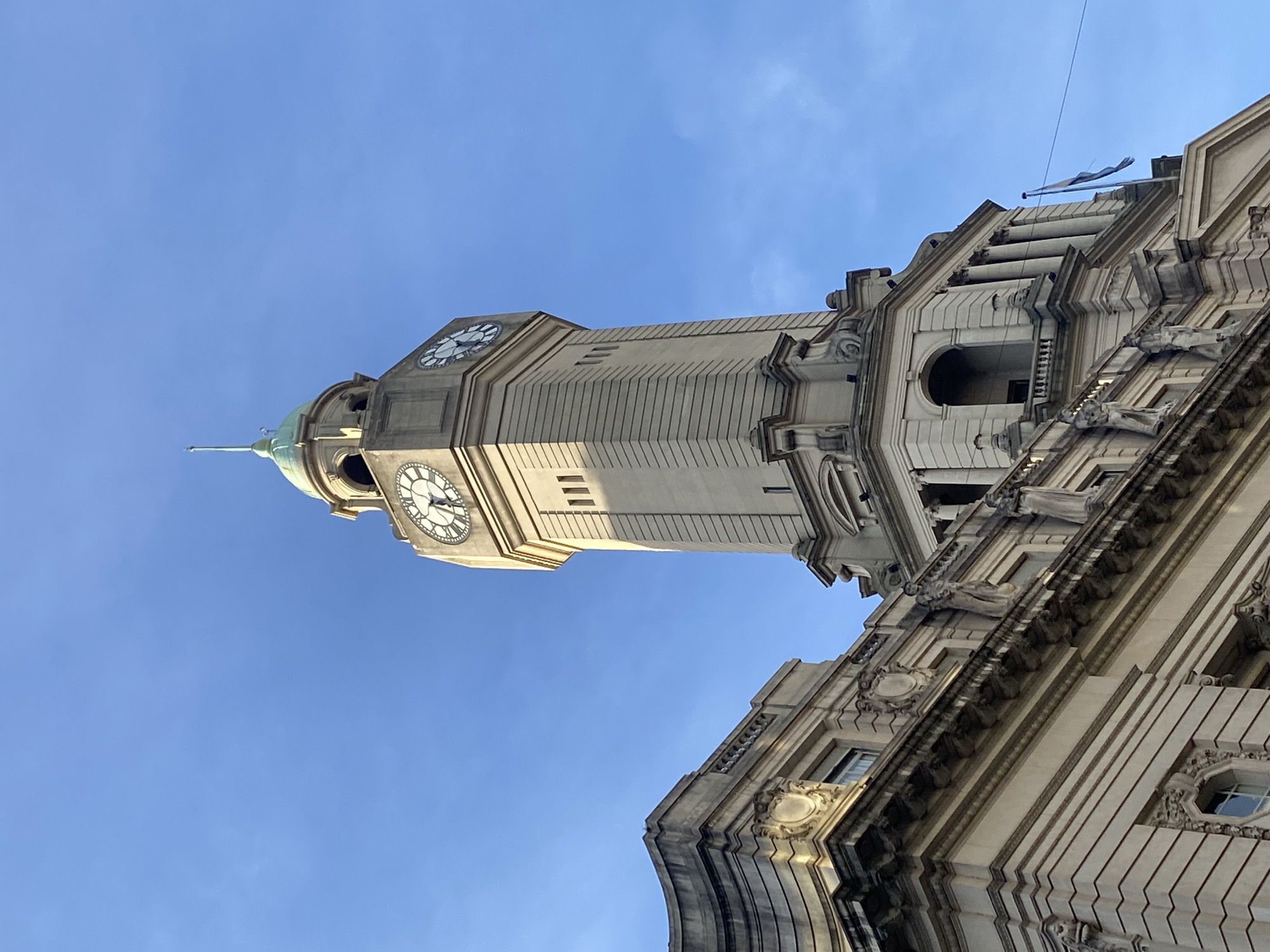 The image captures a low-angle view of the Torre Monumental, also known as the English Clock Tower, situated in the Retiro neighborhood of Buenos Aires, Argentina. The clock tower is prominently featured, with its clock faces visible against a clear blue sky. Adjacent to the clock tower is a portion of a building exhibiting ornate architectural details. The building's facade includes classical elements, such as columns, arches, and decorative carvings. The sky is a solid blue with no clouds. A flag is barely visible on the building to the right of the clock tower.