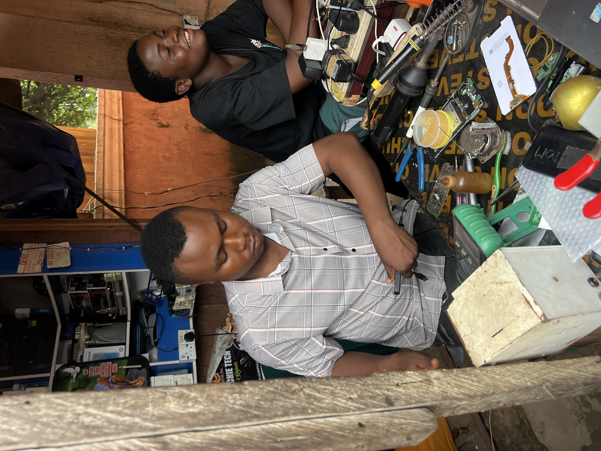 In Nkpor, Nigeria, two individuals are present in a dimly lit workshop. The man in the foreground, wearing a checkered shirt, is seated and appears to be focused on working with electronic components, holding a small tool in his right hand. To his right, a young person in a black t-shirt is looking away from the camera with their eyes closed and a smile on their face, suggesting a moment of amusement or relaxation. The workshop environment is cluttered with various electronic devices and tools, including circuit boards, wires, a soldering iron, and a power strip filled with plugs and adapters. A donation box is visible on the left side of the frame. The background shows shelves filled with more electronic equipment and supplies. The overall scene depicts a busy but organized workspace where electronic repairs or assembly are likely taking place.
