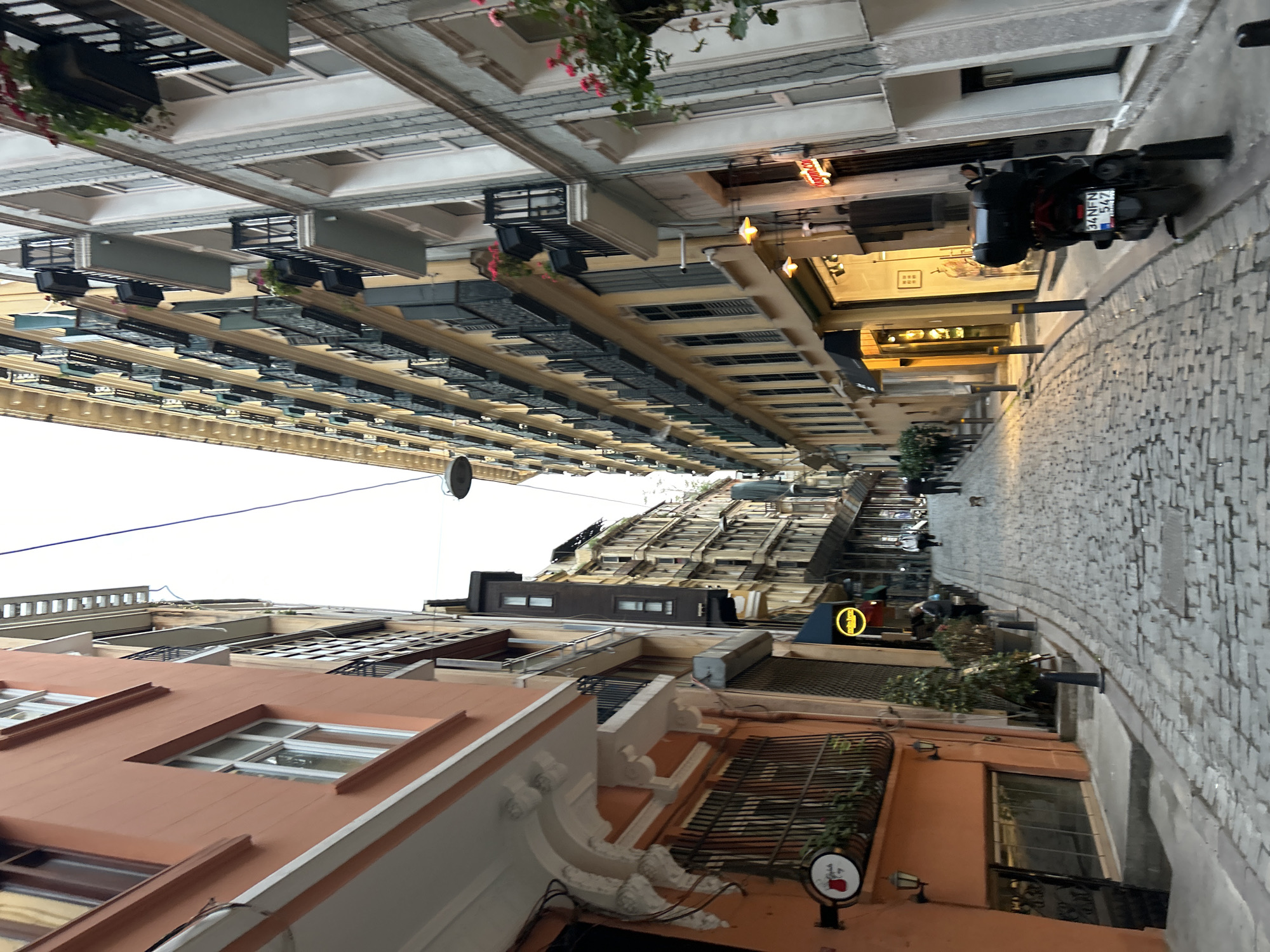The image captures a street scene in Beyoğlu, Turkey, viewed from a low angle looking upwards. The narrow street is paved with light gray cobblestones. Buildings with varied architectural styles line both sides, creating a deep perspective. Some buildings are painted in terracotta hues with white trim, while others display cream and gray façades, some with ornate detailing. Multiple windows of different sizes are visible. Numerous air conditioning units are affixed to the buildings. Several hanging flower baskets adorn the exteriors. A motorcycle parked on the right side of the street is in the foreground. The street is lined with shops and restaurants, indicated by signs and awnings. A dark silhouette of people is visible. The sky appears overcast, creating diffused lighting.