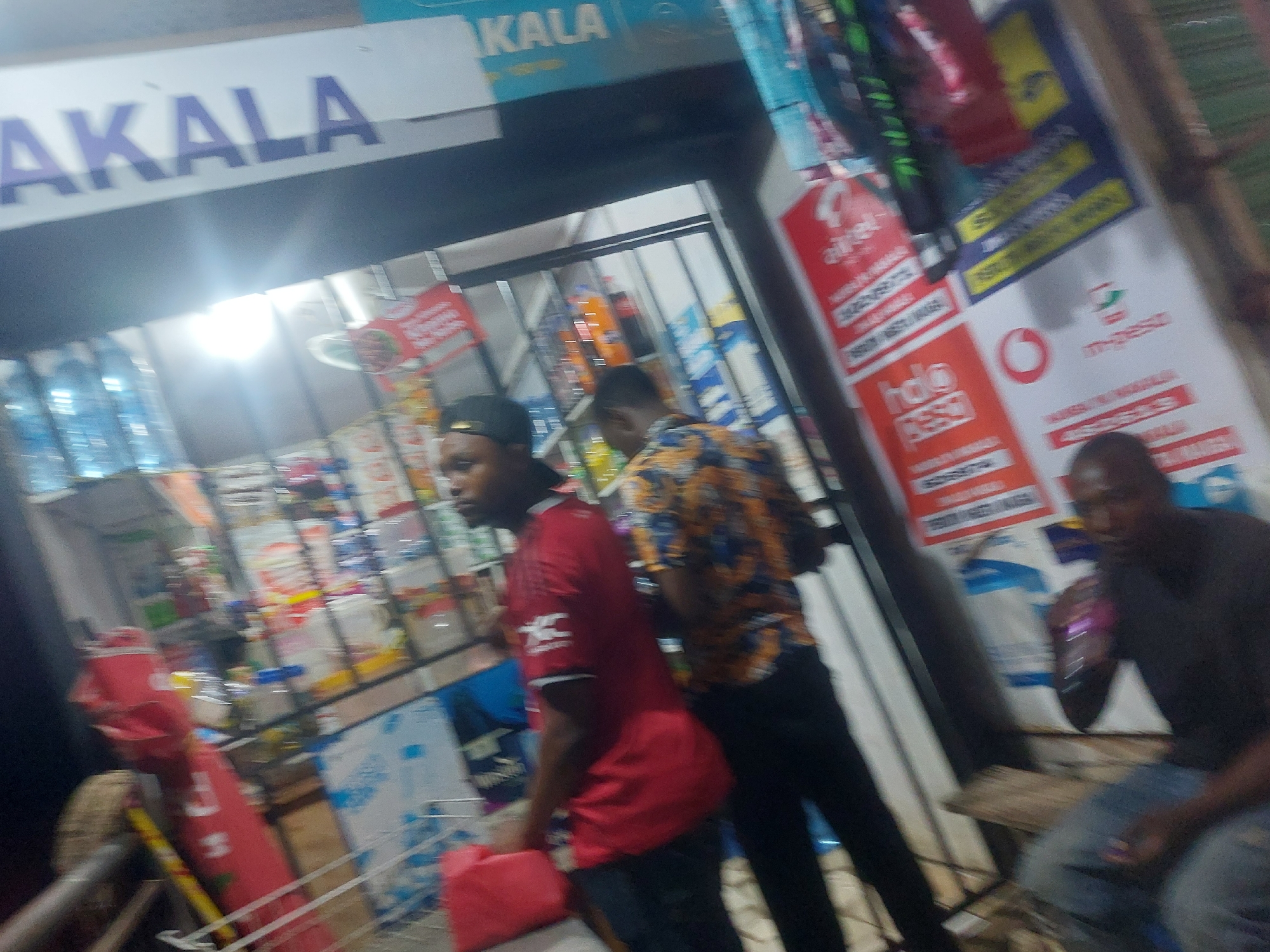 The image shows a small shop in Dar es-Salaam, Tanzania, identifiable by the Swahili word "AKALA" on the shop sign and the presence of mobile money advertisements (e.g., "Halo Pesa," "M-PESA"). The shop is stocked with various goods, including bottled water and snacks. Three men are visible: two are customers, one wearing a red shirt and the other a patterned shirt, while the third man is seated near the shop entrance, looking at his mobile phone. The shop appears to be located on a street, with a simple wooden bench outside. The overall environment suggests a typical urban setting in Dar es-Salaam.