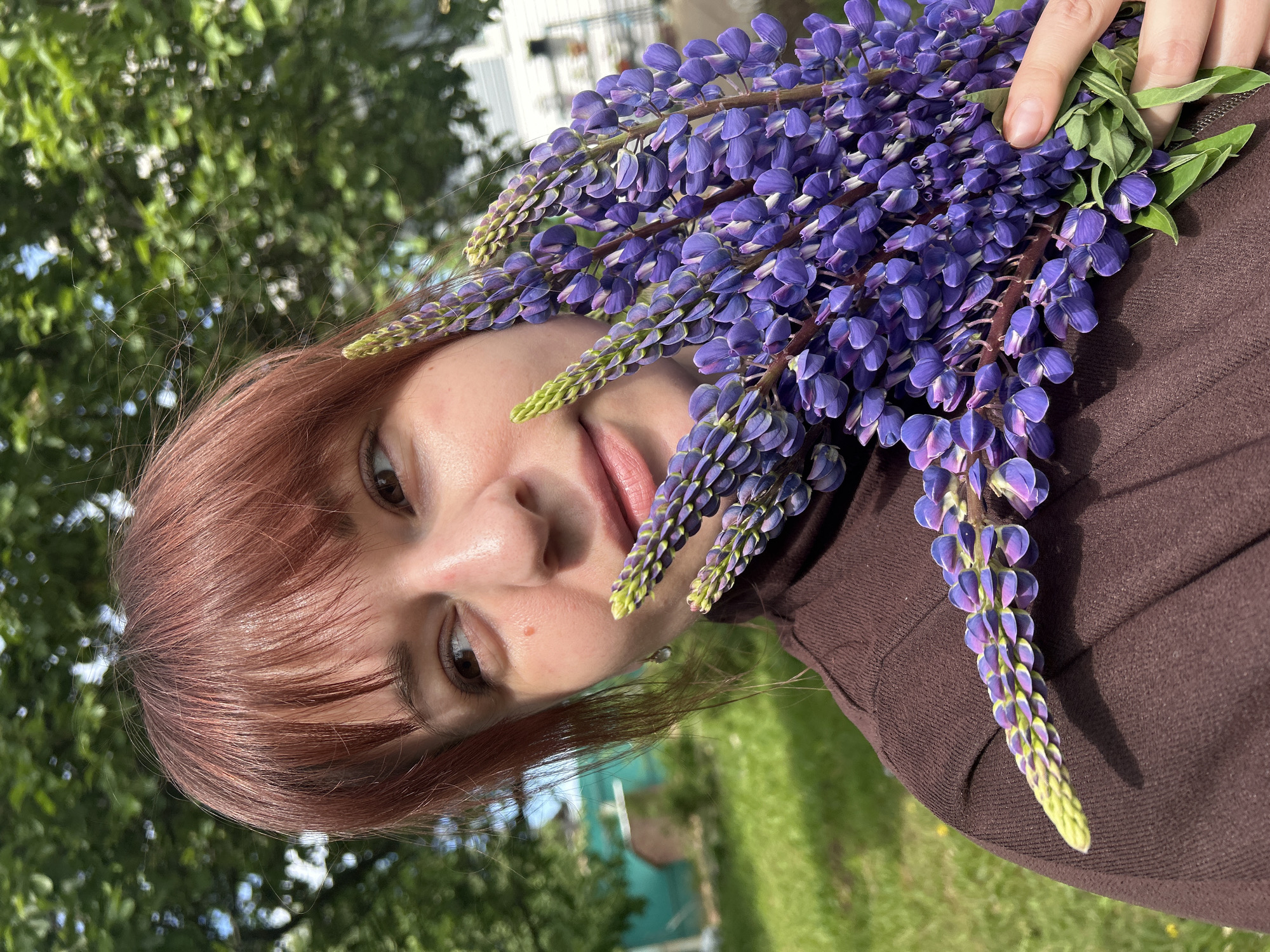 A woman with reddish-brown hair and bangs is holding a bouquet of purple lupine flowers in front of her face. She is wearing a brown turtleneck sweater. The background consists of green trees and grass, with a building visible in the distance. The photograph appears to have been taken outdoors. The location given is Фоки, Russia, but there is no way to verify this information from the image itself.