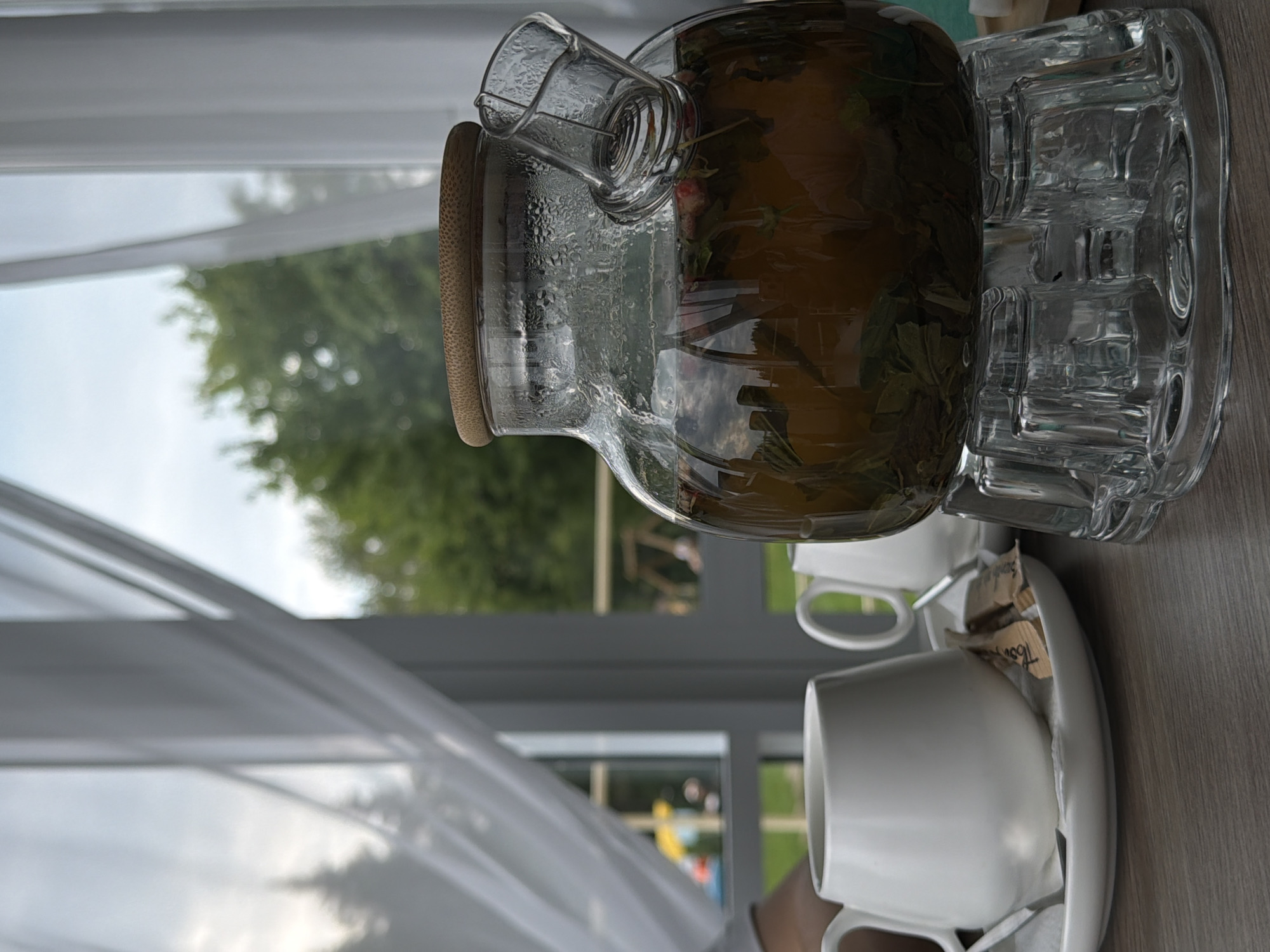 This image depicts a glass teapot filled with tea and leaves, resting on a clear glass stand. Beside the teapot, two white ceramic mugs are placed on saucers, with packets of sugar on one of the saucers. The scene is set indoors, with white curtains partially obscuring a window that looks out onto a green, leafy exterior. The overall ambiance suggests a relaxed setting, possibly a cafe or a home dining area. The location is specified as Chusovoy, Russia.