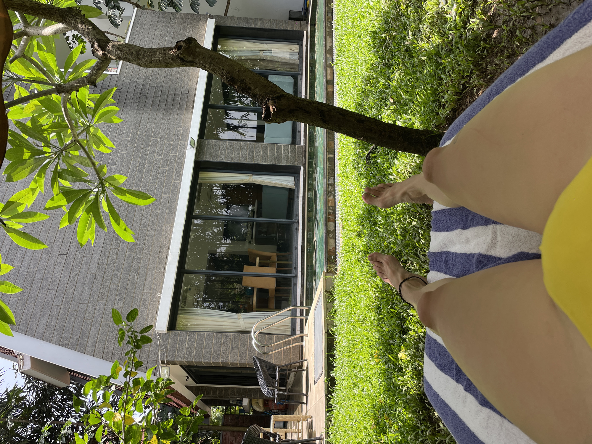 In the city of Badem, India, a person lies on a striped lounge chair in a grassy area. Their legs, covered by a yellow garment at the bottom, are extended towards a swimming pool. The pool is located in front of a modern building with a stone facade and large glass windows. A tree with green leaves stands to the right of the frame, partially obscuring the view of the building. The scene appears to be a tranquil outdoor setting, possibly a resort or private residence.
