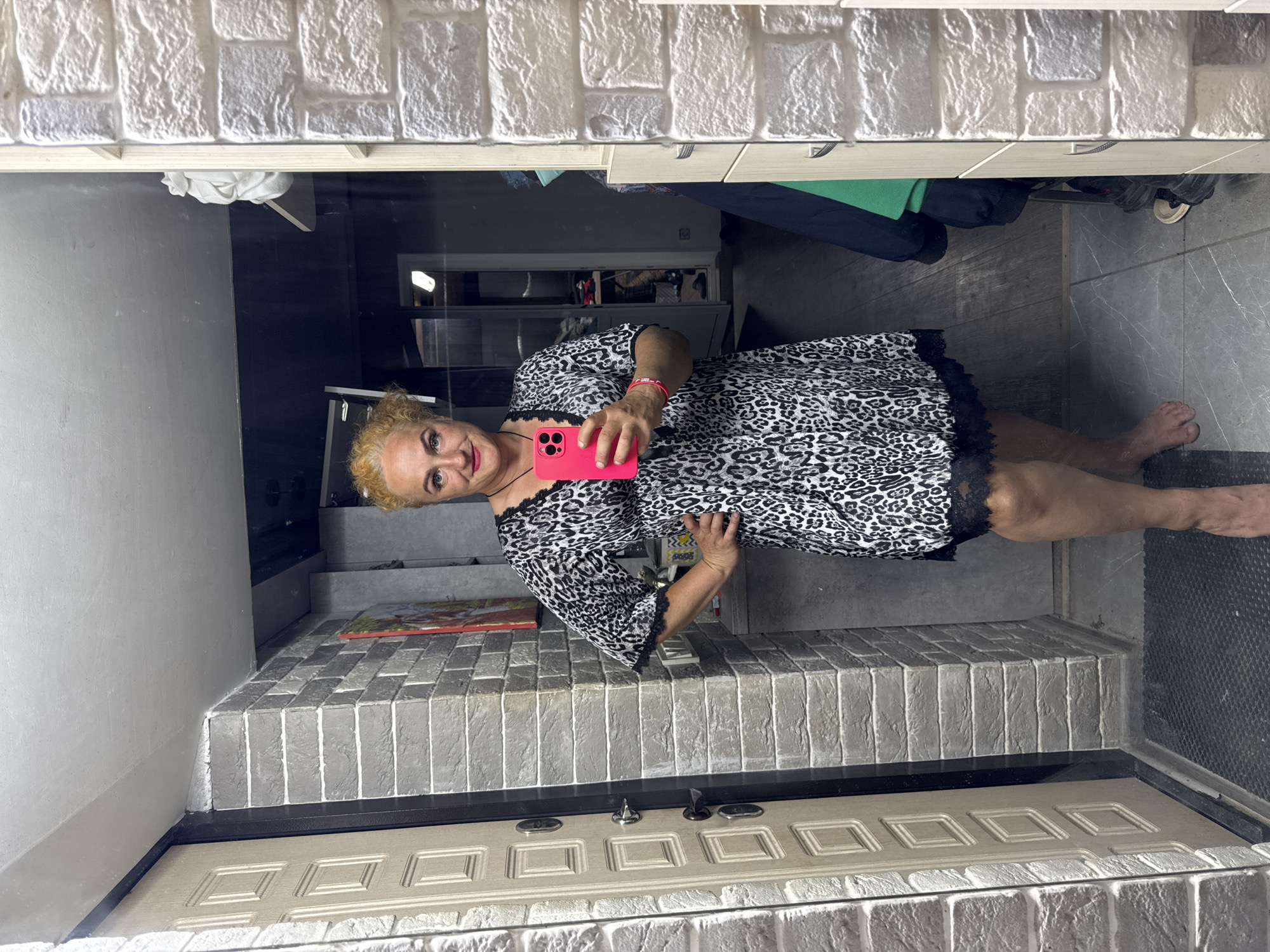 A woman is taking a mirror selfie in what appears to be a hallway or entryway. She has blonde hair and is wearing a leopard print dress with black lace trim. She is holding a pink smartphone in her right hand and her left hand is on her hip. The background includes a door on the left, a wall made of grey and white textured bricks, and a closet or cabinets on the right. There is also a floor mat and a wooden floor visible. The image does not contain enough information to determine its location in the city of Chaikovsky, Russia.