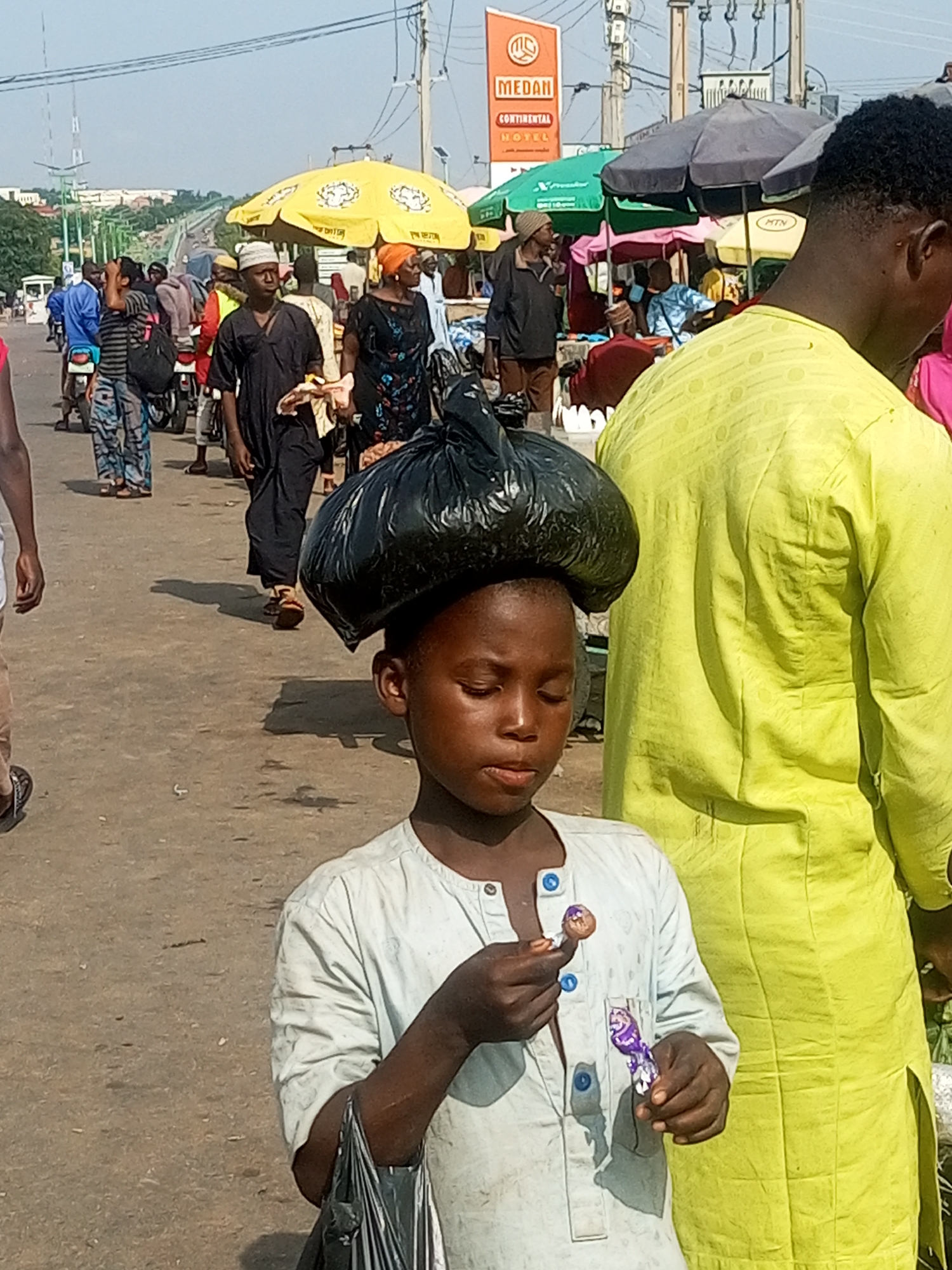 In a bustling street scene in Tempure, Nigeria, a young boy, the primary subject, walks with a large black plastic bag balanced precariously on his head. He is wearing a light blue, buttoned shirt. In his right hand, he holds a lollipop, examining it with his eyes closed. Another wrapped candy or snack is visible in the same hand. Behind him, an adult wearing a bright yellow shirt stands prominently, their back to the camera. The background reveals a busy marketplace with several stalls and umbrellas, including a prominent yellow umbrella and a green and black umbrella. A sign for "MEDAN CONTINENTAL HOTEL" is visible in the distance, indicating the location. Other people, some carrying bags or riding motorcycles, are also present, navigating the crowded street. The ground is a paved road, suggesting an urban environment. The sky is clear and bright.