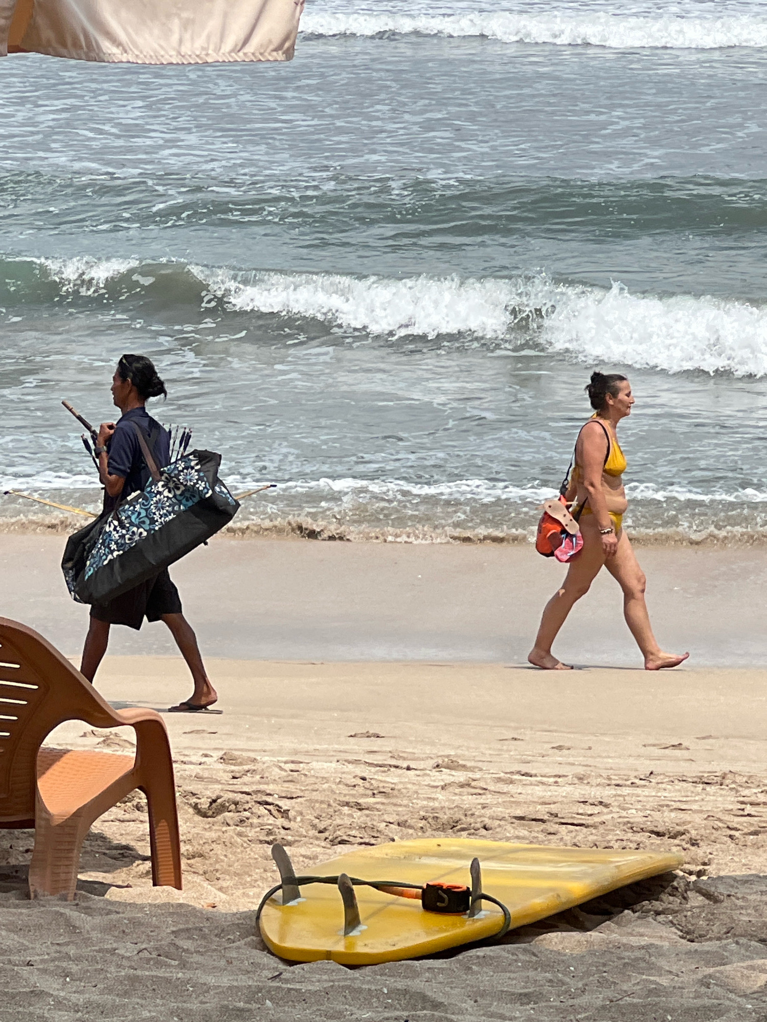 In Kuta, Indonesia, on a sandy beach with the ocean in the background, two individuals are walking along the shoreline. The person on the left, a male with his hair tied back, wears a dark blue t-shirt and black shorts, carrying a large patterned bag slung over his shoulder. He also carries a long, thin object, possibly a pole or a staff. To the right, a female wearing a yellow bikini walks by, carrying a bag with red and orange accents over her shoulder. In the foreground, a yellow surfboard with fins lies on the sand, and an orange plastic chair is partially visible. The waves are breaking gently on the shore.