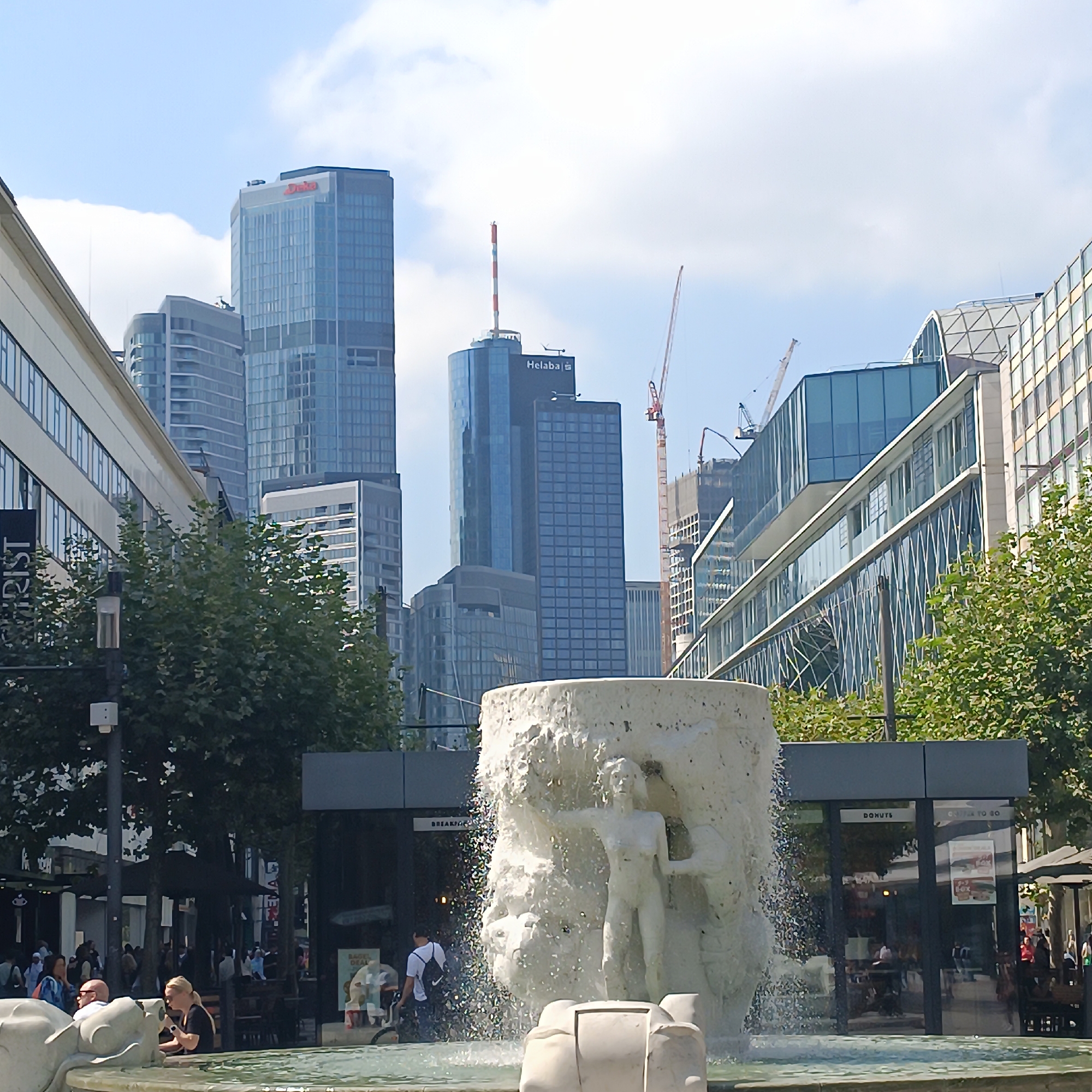 The image depicts a public square in Frankfurt, Germany, dominated by modern skyscrapers and a fountain. In the foreground, a large, textured concrete fountain features sculpted figures, with water spraying from its top. To the left of the fountain, people are visible, some sitting and others walking. A street lamp stands nearby. Behind the fountain and extending into the midground, several contemporary buildings are prominent. Notably, a tall, blue-glass skyscraper with "Helaba" branding on its upper section is visible, as well as other high-rise structures made of glass and concrete. Construction cranes are also present, suggesting ongoing development. Trees and greenery are interspersed throughout the scene, particularly on the left side of the image, softening the urban landscape. The sky is blue with scattered clouds, indicating a daytime setting. The overall impression is of a bustling urban environment with a mix of contemporary architecture and public amenities.