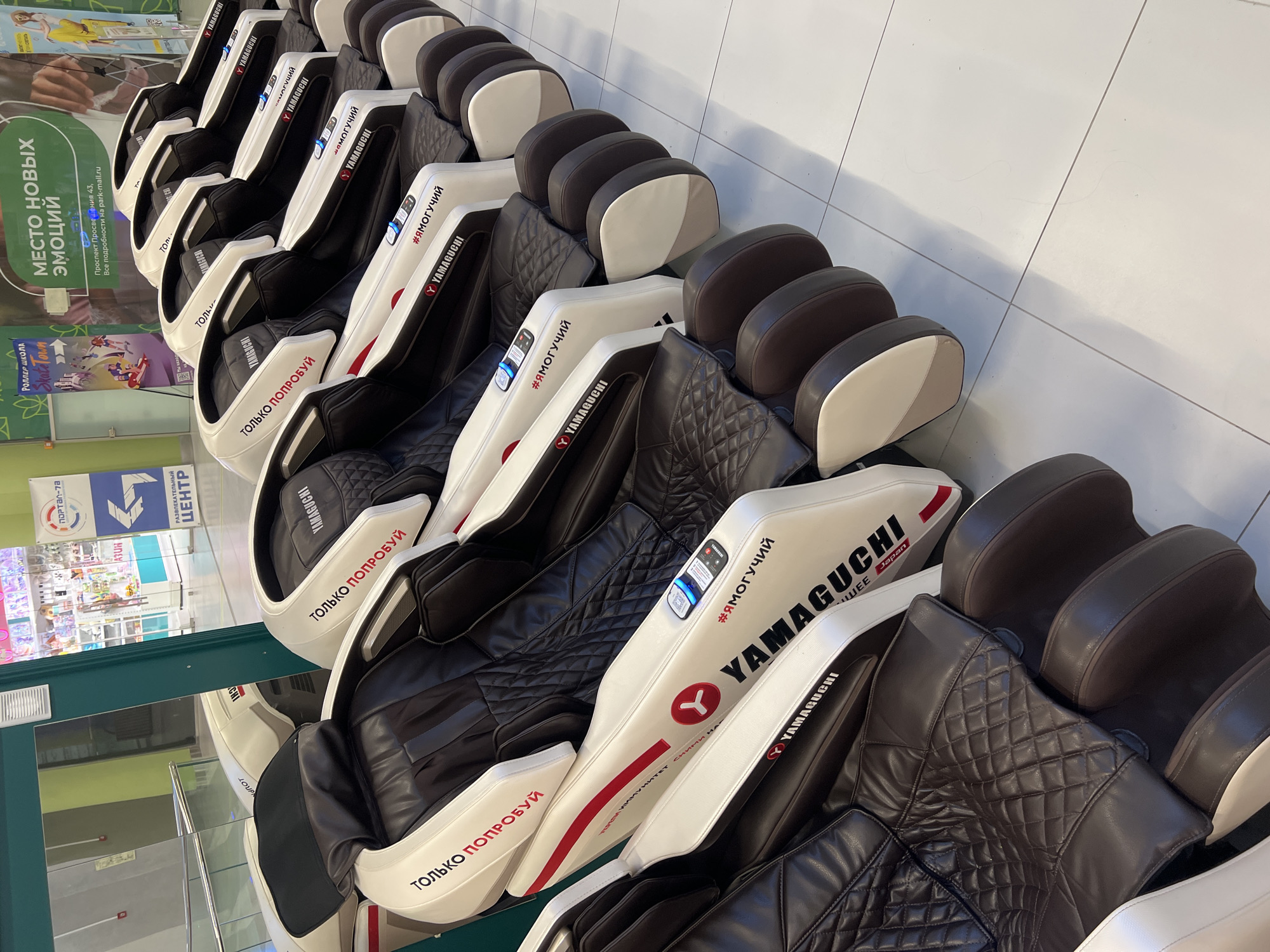 The image shows a row of Yamaguchi massage chairs displayed in a retail setting, likely a shopping mall in Irinovka, Russia. The chairs are predominantly white with black leather upholstery and dark brown accents. Several chairs have the "YAMAGUCHI" logo and the Russian text "#ЯМОГУЧИЙ" (meaning "I am powerful" or "I can") visible. One chair also displays the text "ТОЛЬКО ПОПРОБУЙ" (meaning "Just try it").
In the background, a large banner with the text "МЕСТО НОВЫХ ЭМОЦИЙ" (meaning "Place of new emotions") and "РОЛЛЕР ШКОЛА Skak Town" (Roller School Skak Town) is visible, along with promotional material for a roller school and a shopping center. There are also shelves stocked with merchandise in the distant background, suggesting a commercial environment. The lighting suggests an indoor setting, likely during daytime hours. No people are visible in the image, but the arrangement of the chairs implies they are on display for customers to try. The overall scene conveys a sense of commercial display and promotion of relaxation and wellness products.