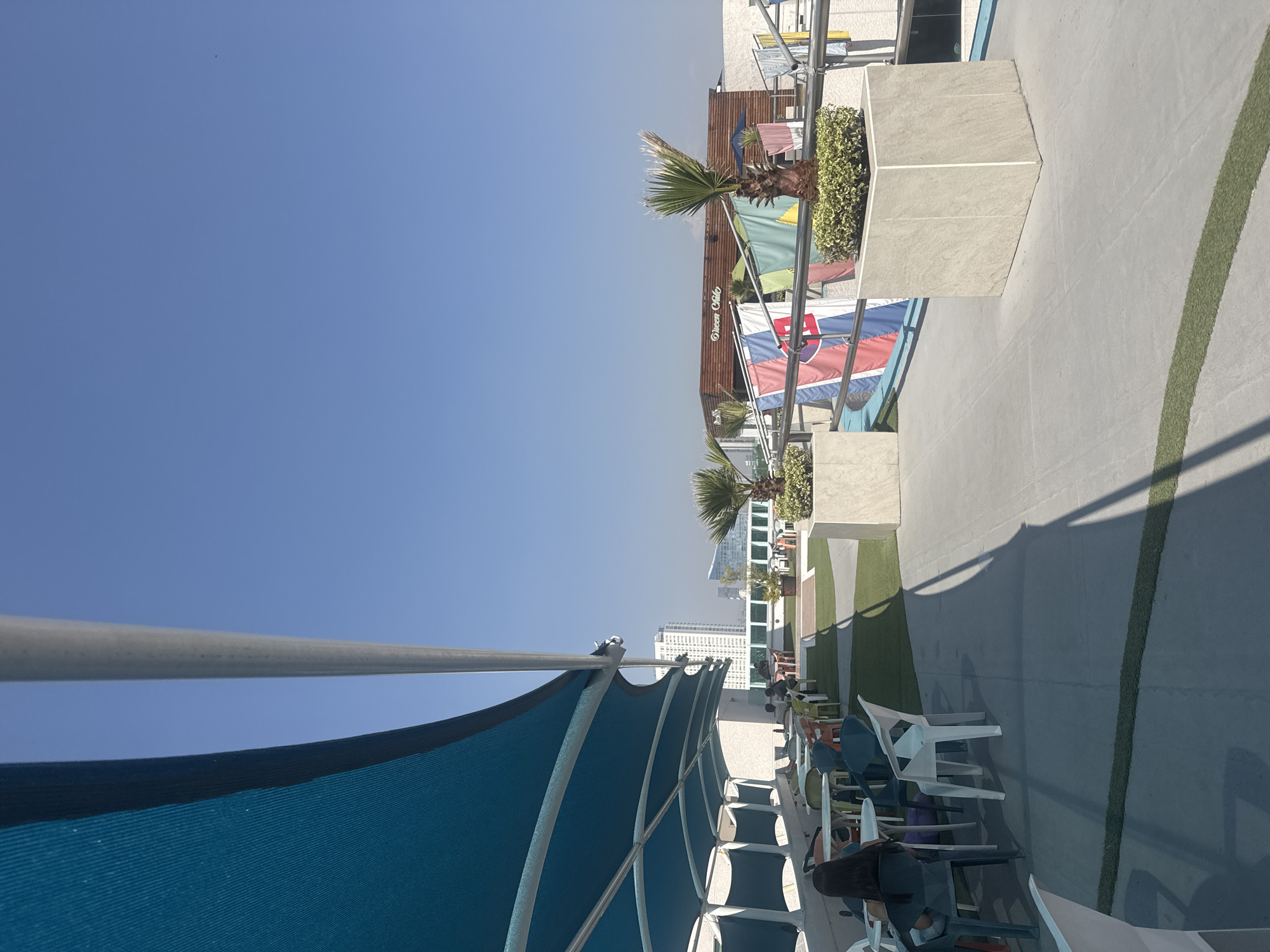 The image shows a rooftop space in Puebla, Mexico. The sky is clear and blue. In the foreground, there's a blue shade structure with metal supports over a seating area. The seating area is populated with various colored plastic chairs arranged in rows. In the middle ground, there's a pathway with artificial grass strips, edged by planters containing small palm trees and greenery. The flags of different countries are displayed alongside a building with "Ocean Club" written on it. In the background, there are tall buildings and structures, indicating a cityscape.