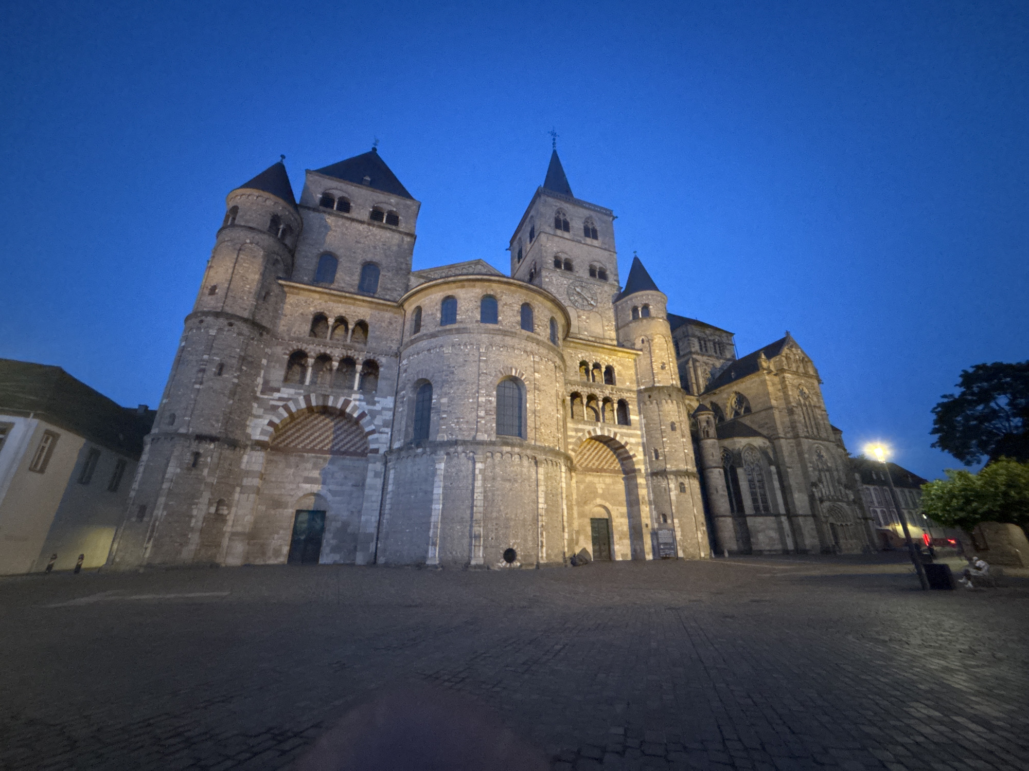 The image shows the Trier Cathedral, also known as the Cathedral of Saint Peter, located in Trier, Germany. The cathedral is a large, ancient Roman Catholic cathedral with multiple towers and a complex architectural design. It is illuminated by artificial lights, highlighting its stone facade. The sky above is a deep blue, indicating that it is dusk or early night. In front of the cathedral is a paved square with cobblestones. There are a few people visible in the distance, including one person sitting on a bench. The overall scene depicts a historic architectural landmark in an urban setting during twilight.